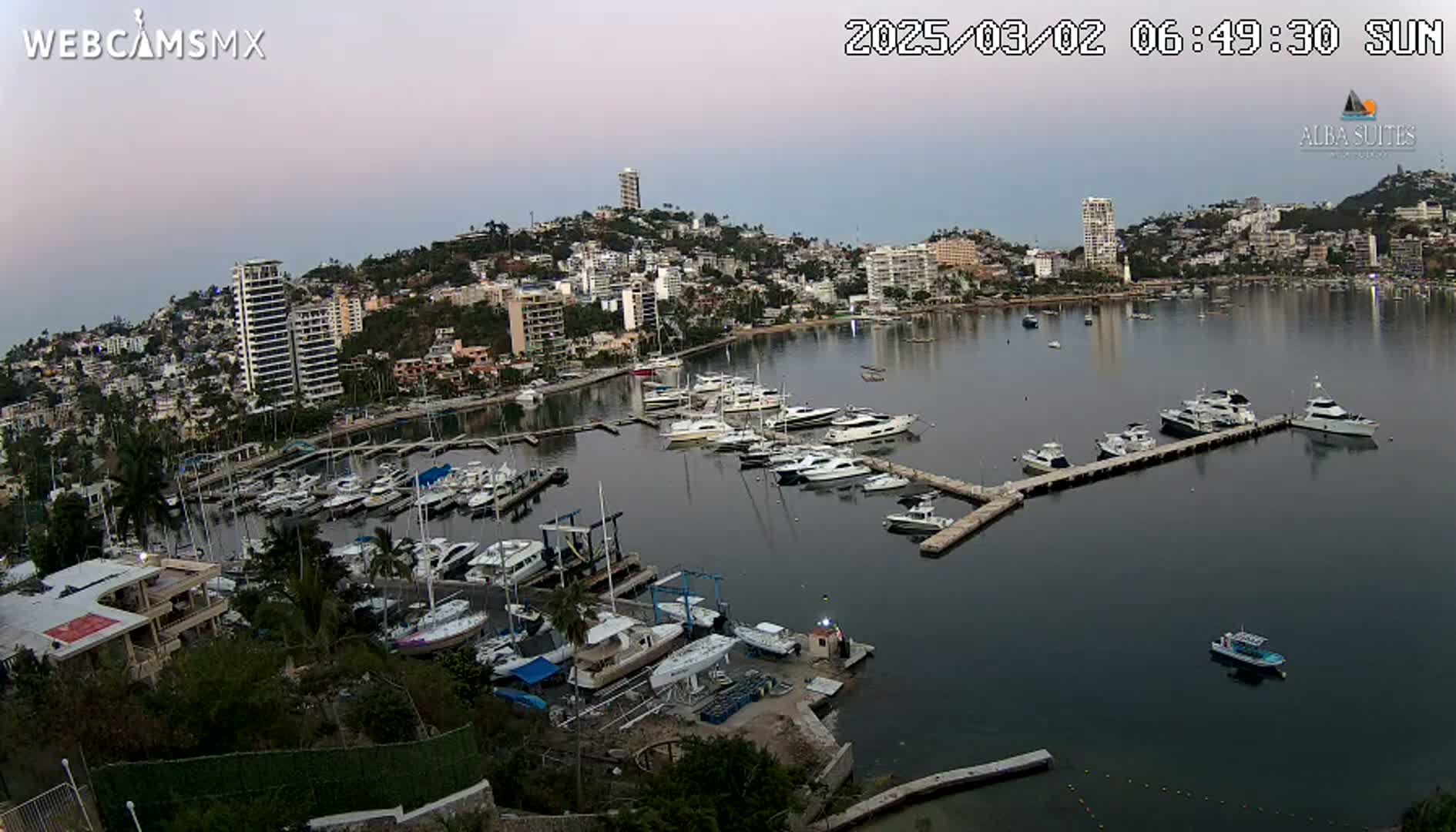 A calm harbor filled with numerous boats and yachts is nestled beside a hillside town under a twilight sky.