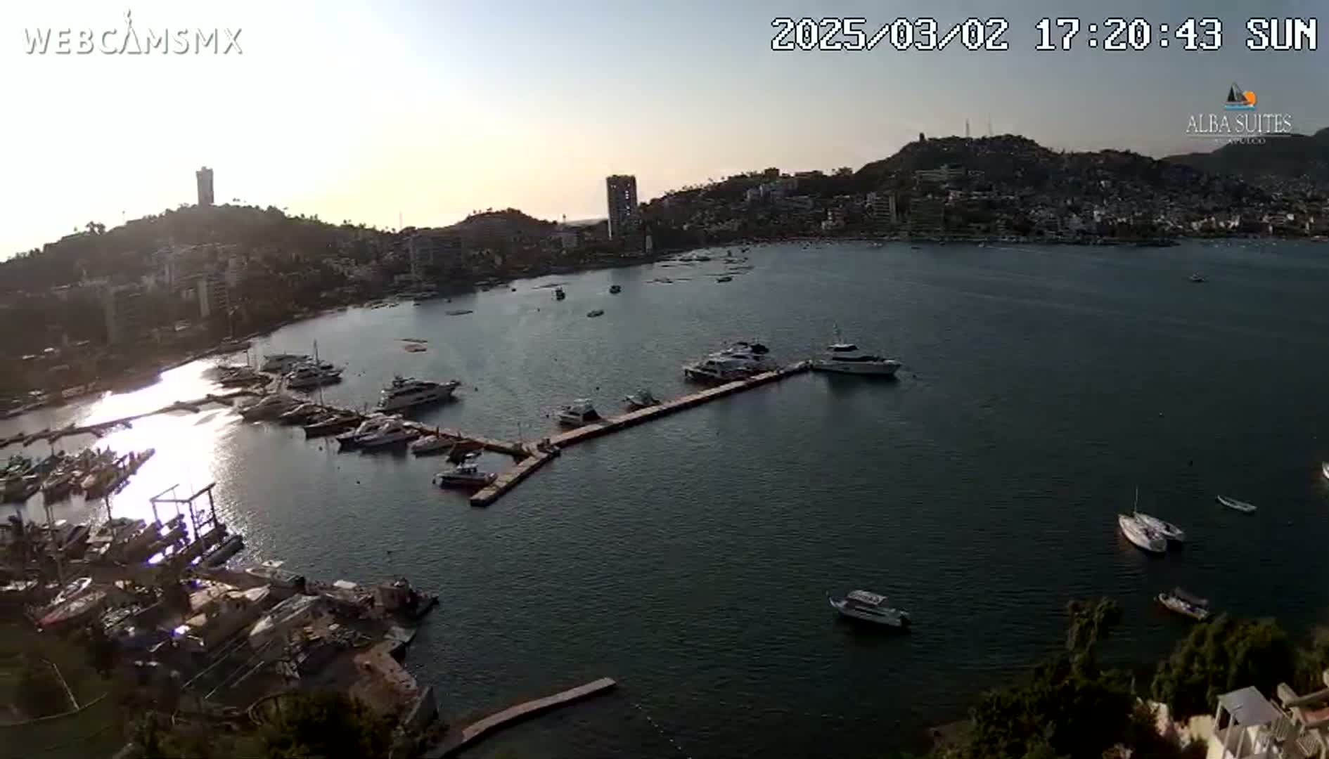 An aerial view shows a calm bay with numerous boats docked at a marina, nestled beside a hillside town under a clear, sunset sky.