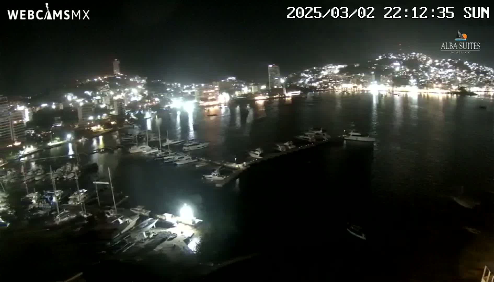 A nighttime aerial view of a city harbor filled with docked boats and illuminated buildings along a calm body of water.