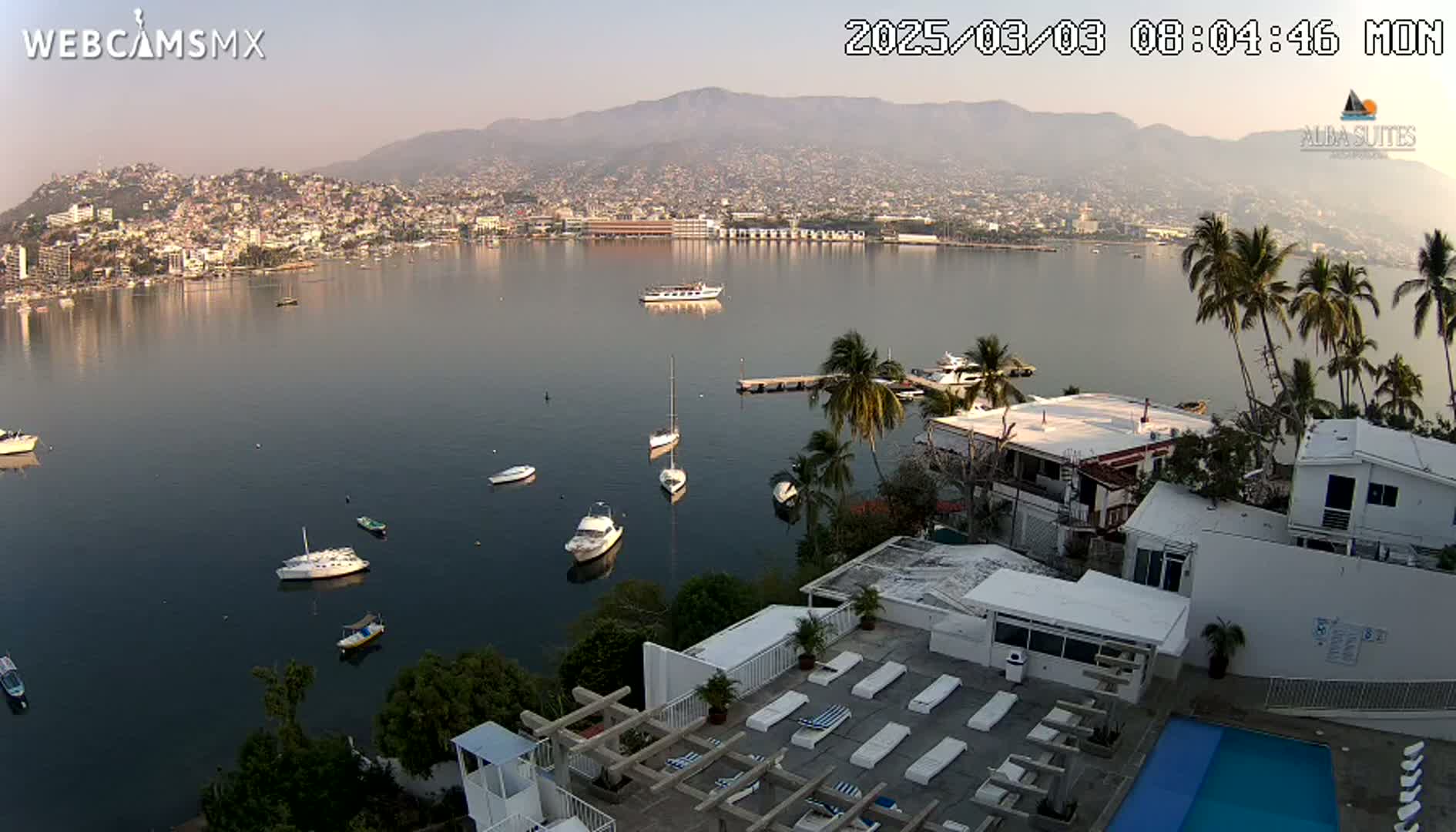 A hazy morning view overlooking a calm bay with numerous boats, a city nestled against mountains in the background, and a white building with a pool and sun loungers in the foreground.