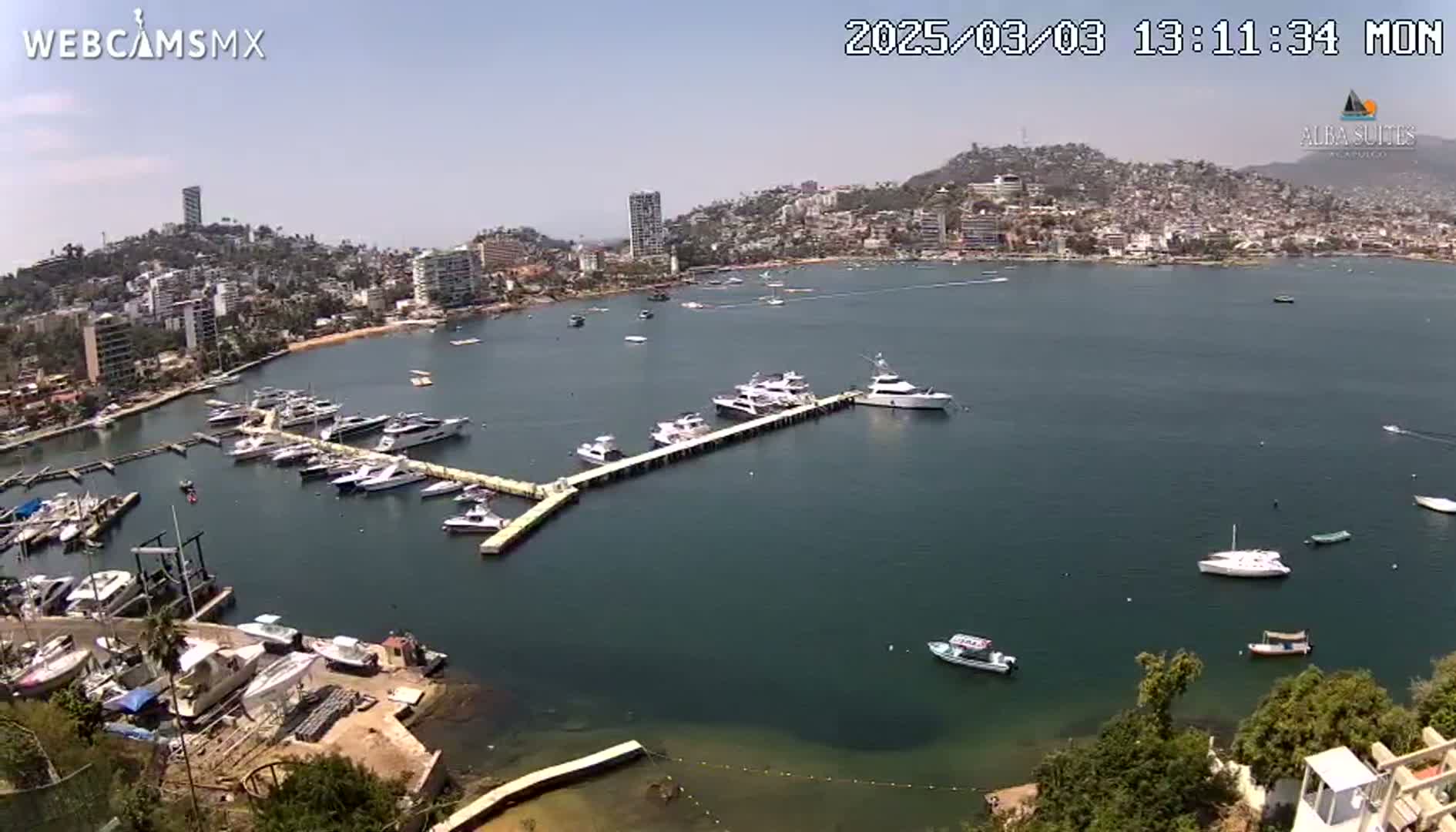 A marina filled with numerous boats and yachts is situated in a calm bay next to a hillside town under a sunny sky.