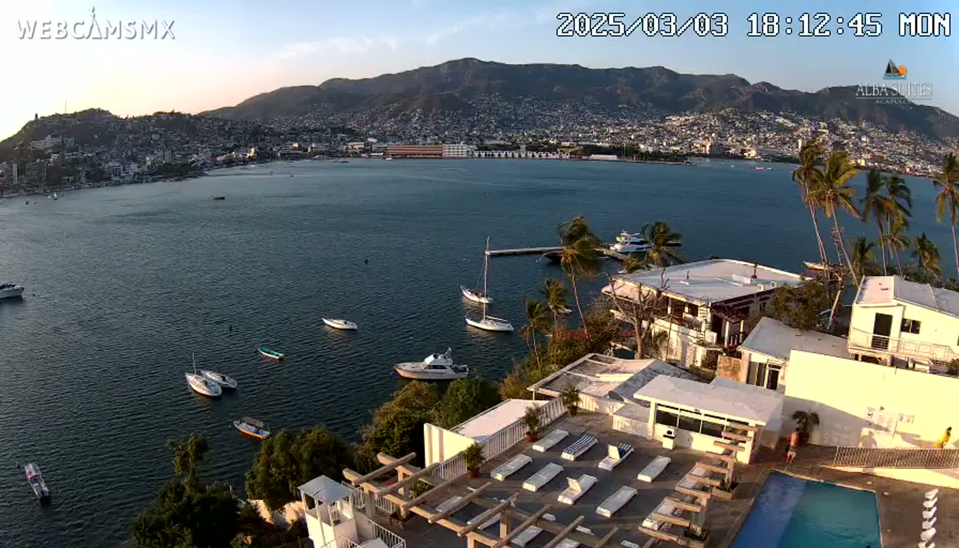 An aerial view shows a calm bay with several boats, a city nestled against mountains in the background, and a white building with a pool and sun loungers in the foreground under a clear evening sky.