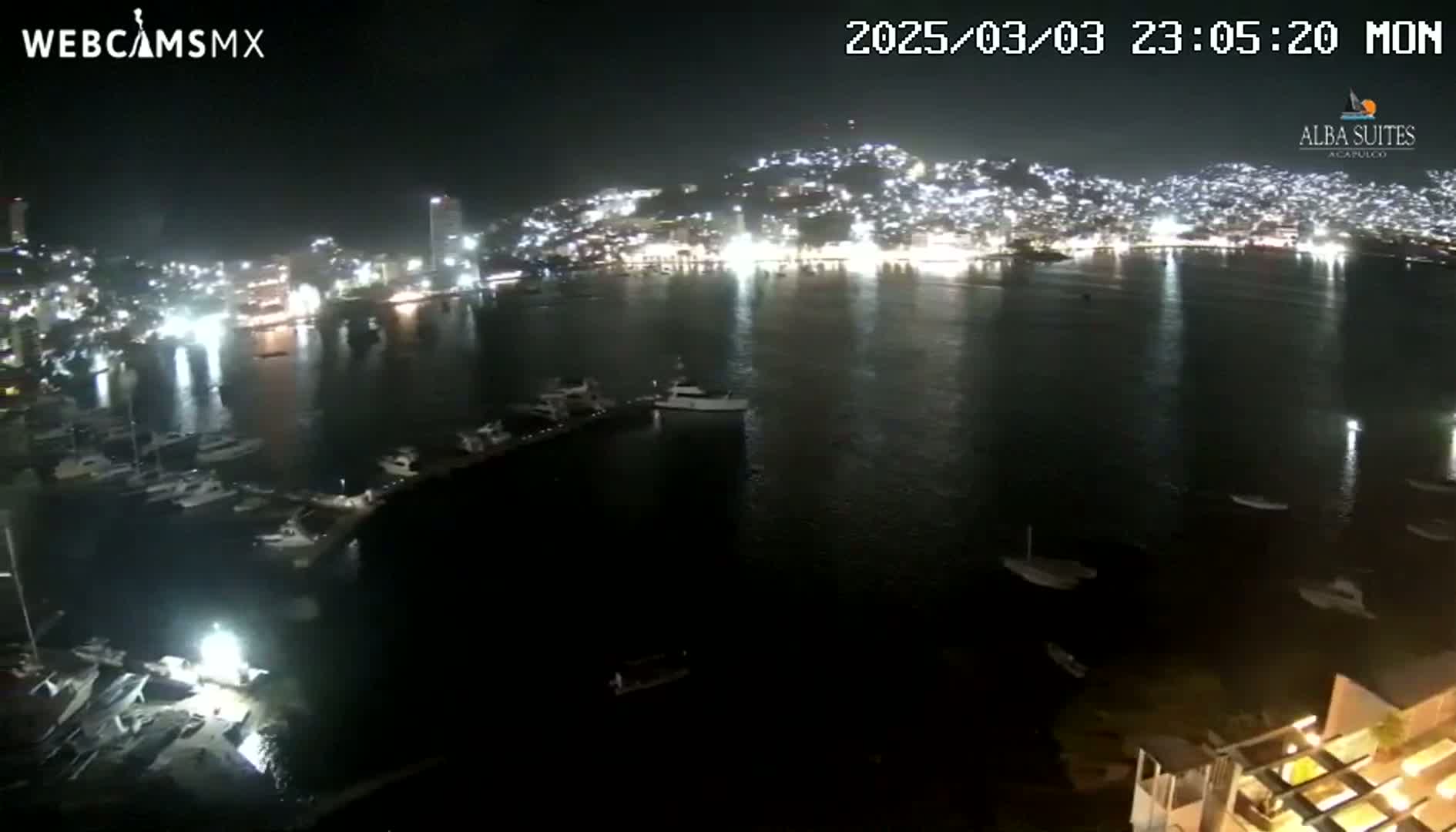 A nighttime view of a brightly lit city across a calm body of water, with numerous boats and yachts in a marina.