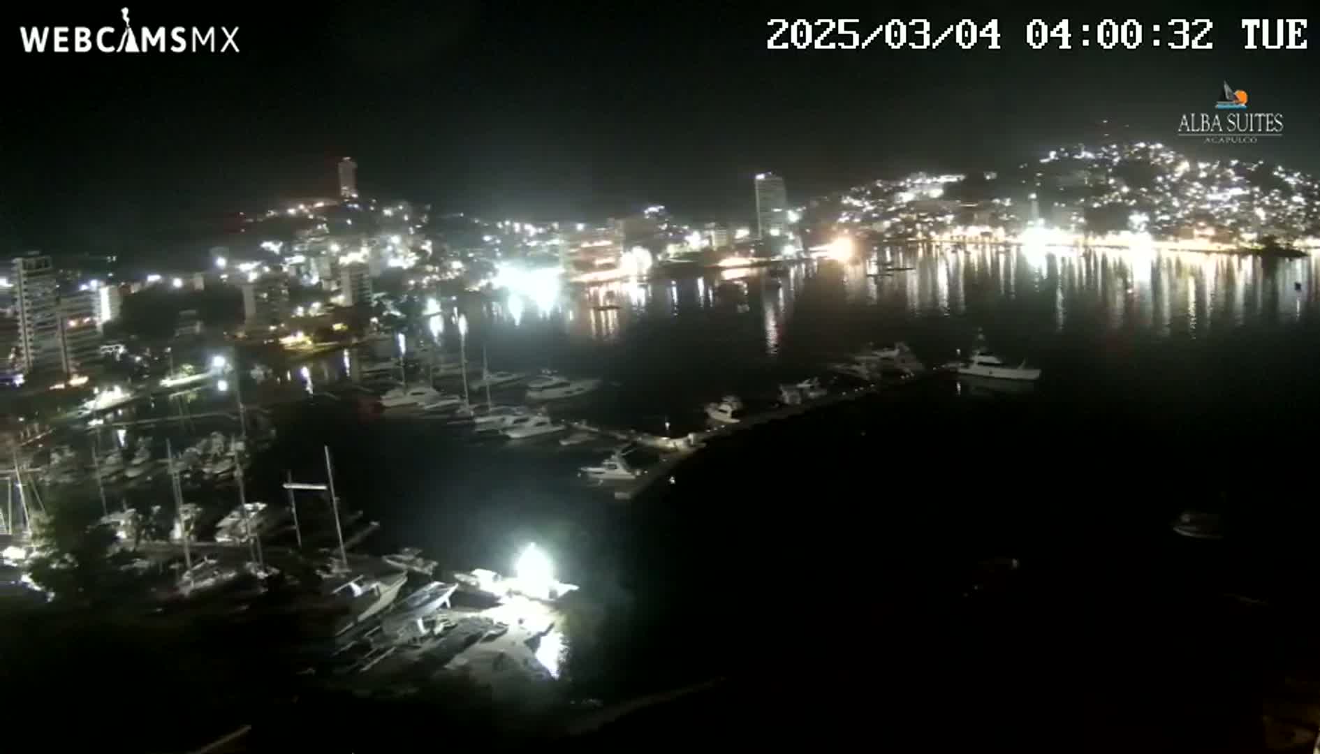 A nighttime aerial view shows a harbor filled with boats and a brightly lit city across the water.