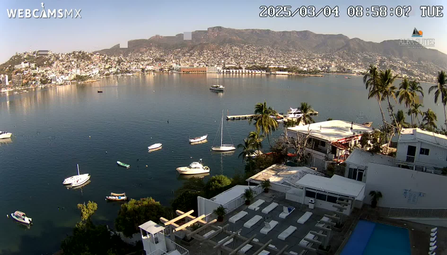 A calm bay with numerous boats, a city nestled against mountains, and a white building with a pool in the foreground under a clear sky.