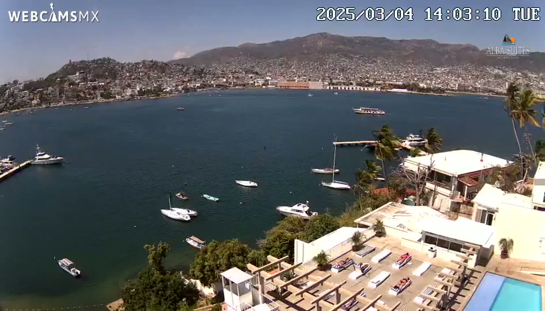 A sunny, coastal city with numerous boats in a bay is viewed from a high vantage point overlooking a building with a swimming pool and sunbathing area.