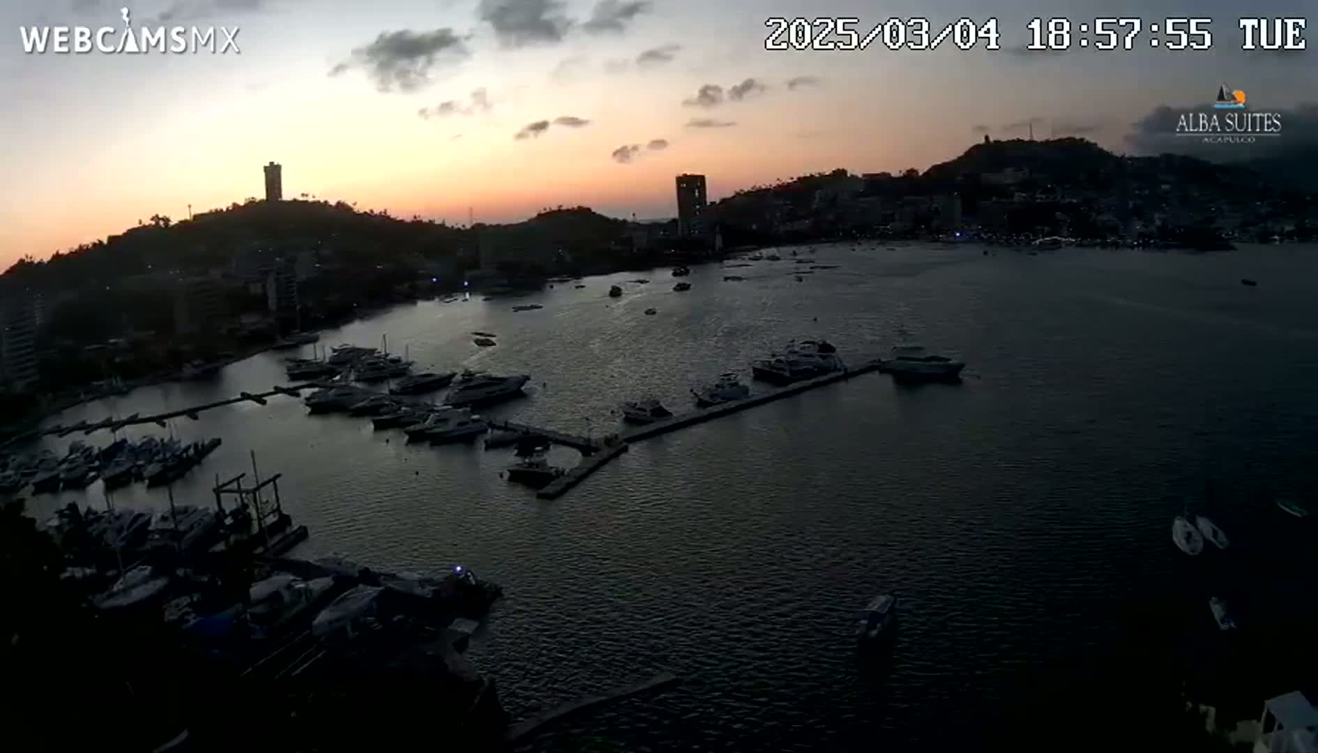 A twilight view shows a harbor filled with numerous boats and yachts, nestled alongside a city silhouetted against an orange and pink sunset sky.