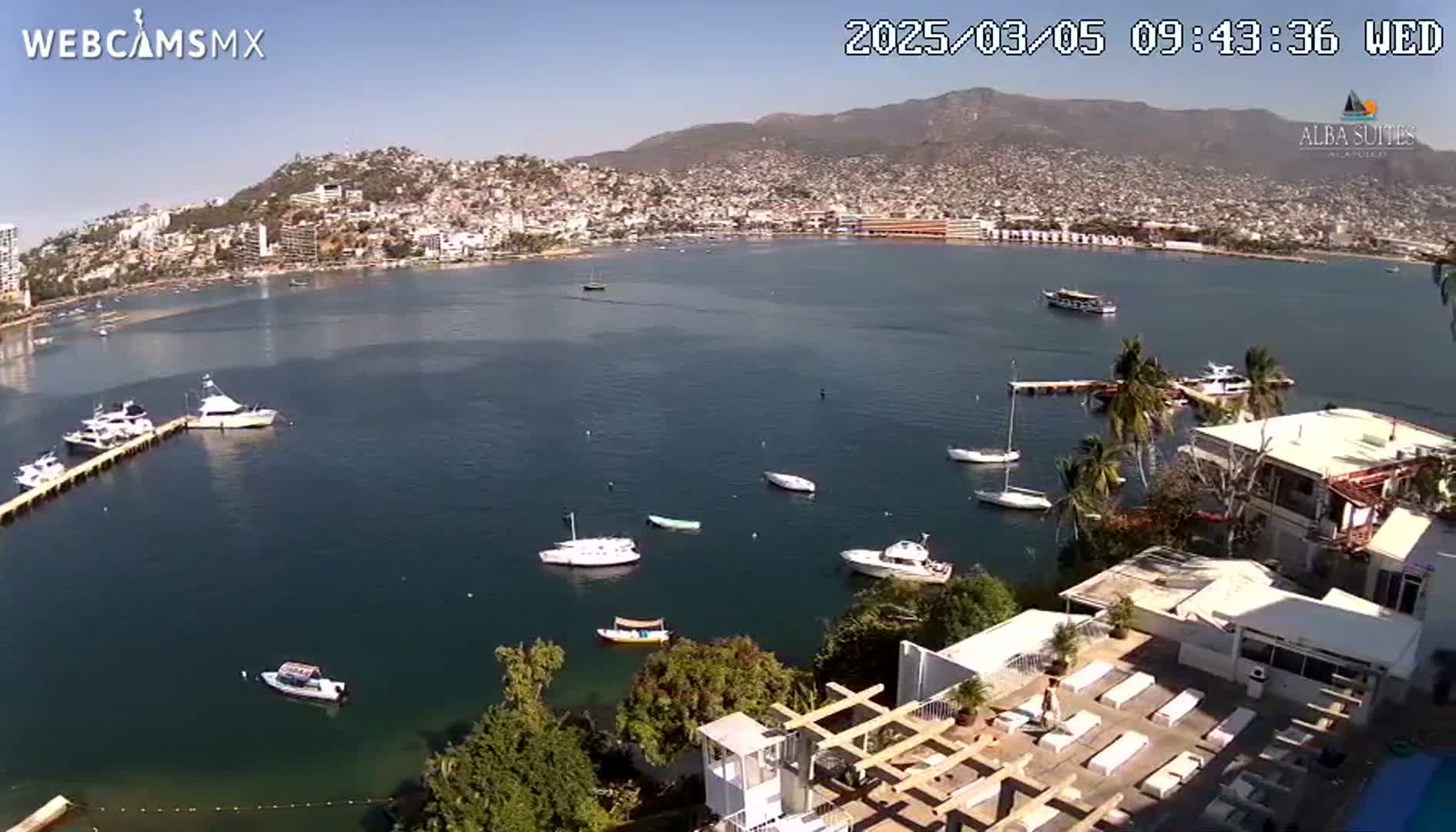 An aerial view of a calm bay with numerous boats, a city nestled against mountains in the background, and a sunny day.