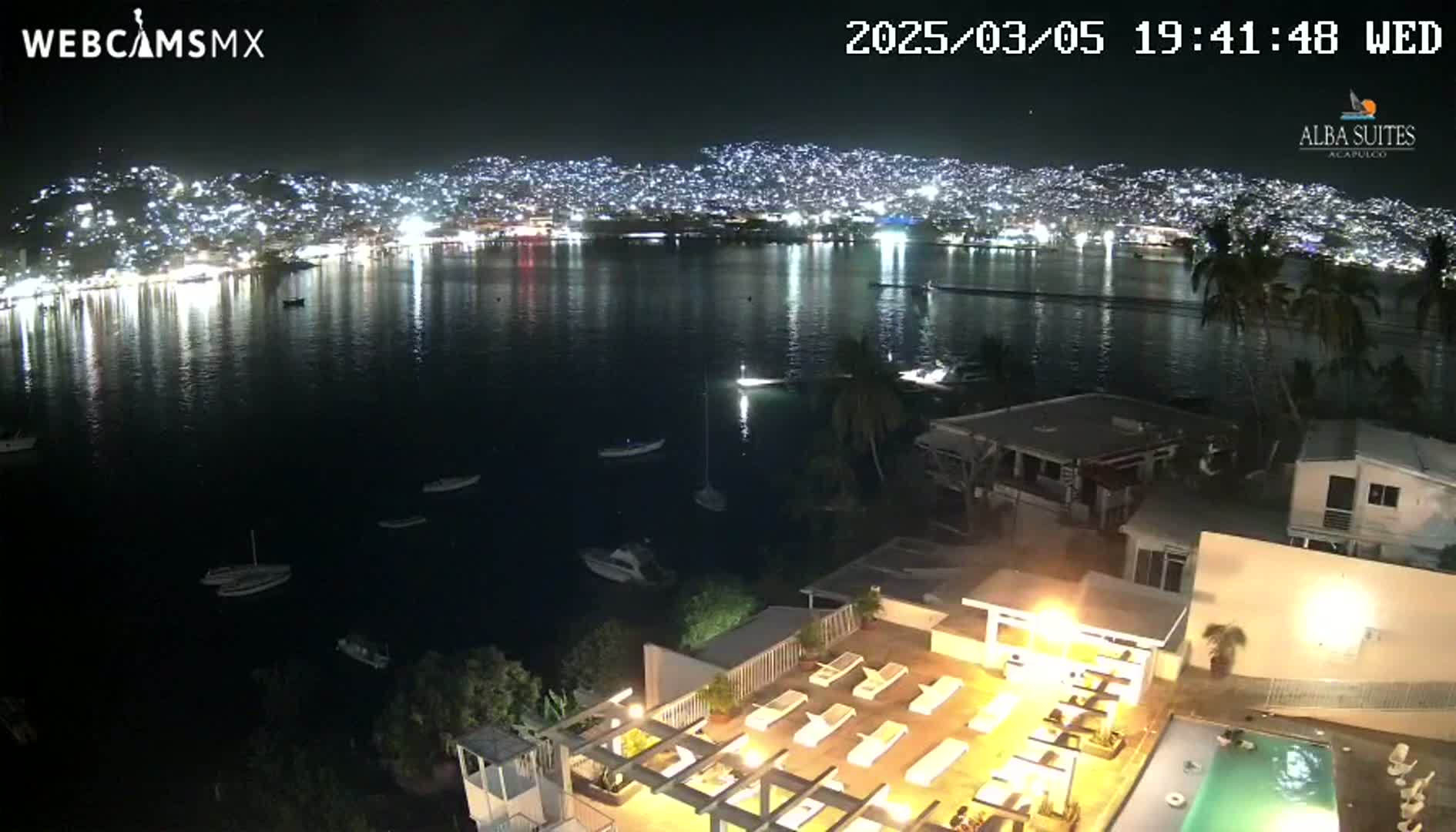 A nighttime view of a city's lights across a calm body of water, with a swimming pool and rooftop deck visible in the foreground.