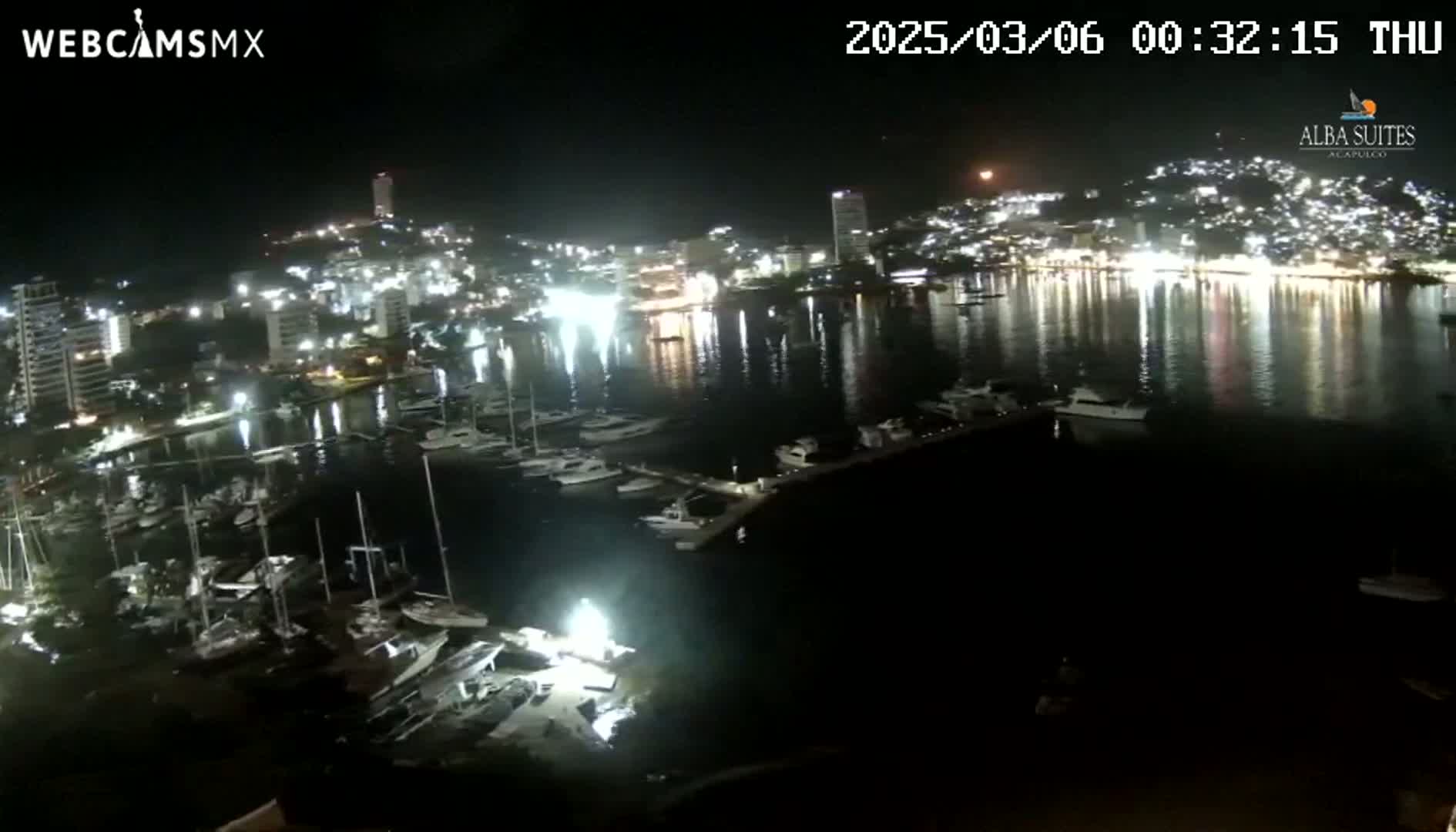 A nighttime aerial view shows a marina filled with boats and a city with brightly lit buildings reflecting in the calm water.