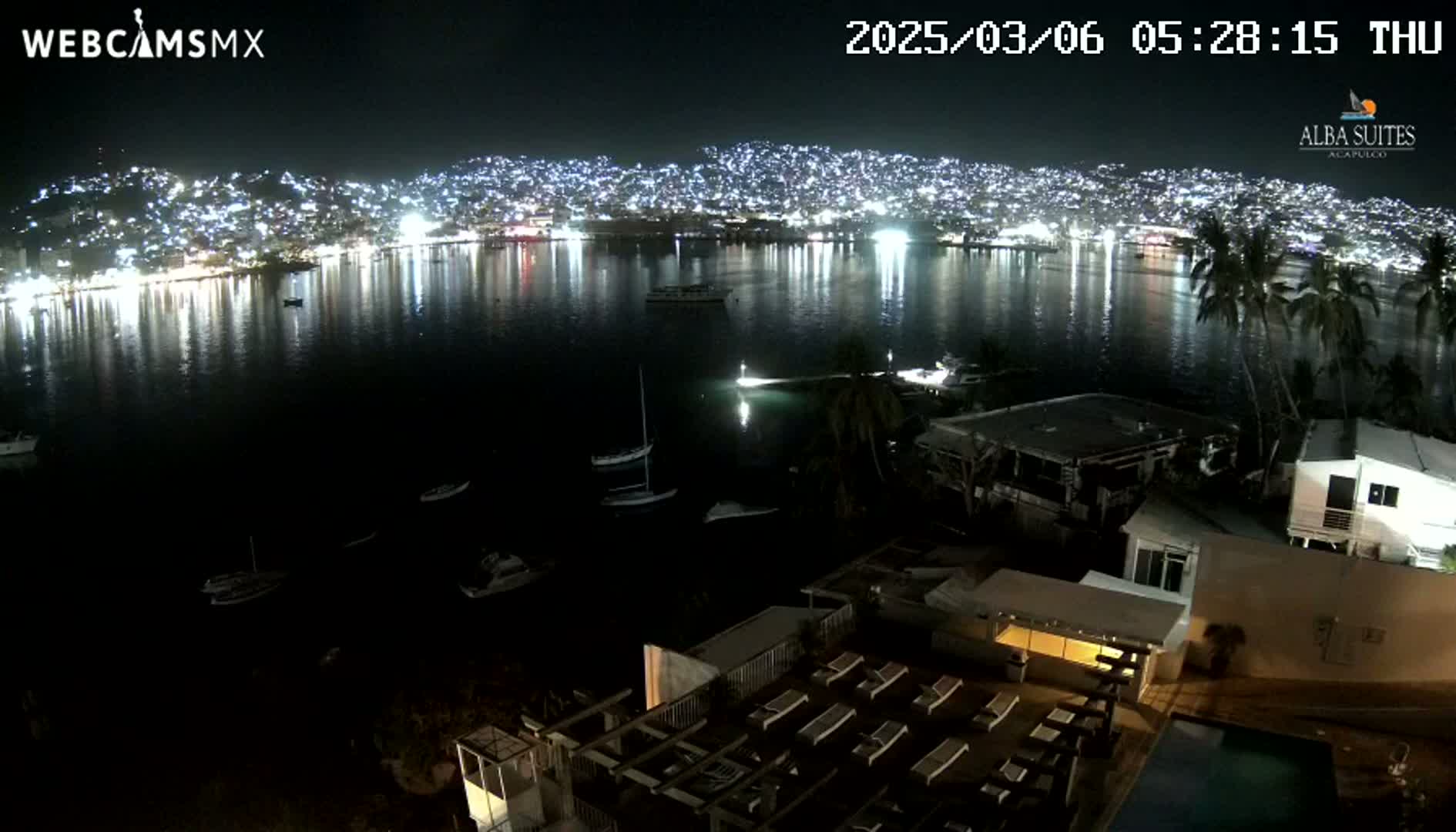 A nighttime view shows a city illuminated across a dark bay, with boats in the water and a building with a pool in the foreground under a clear sky.