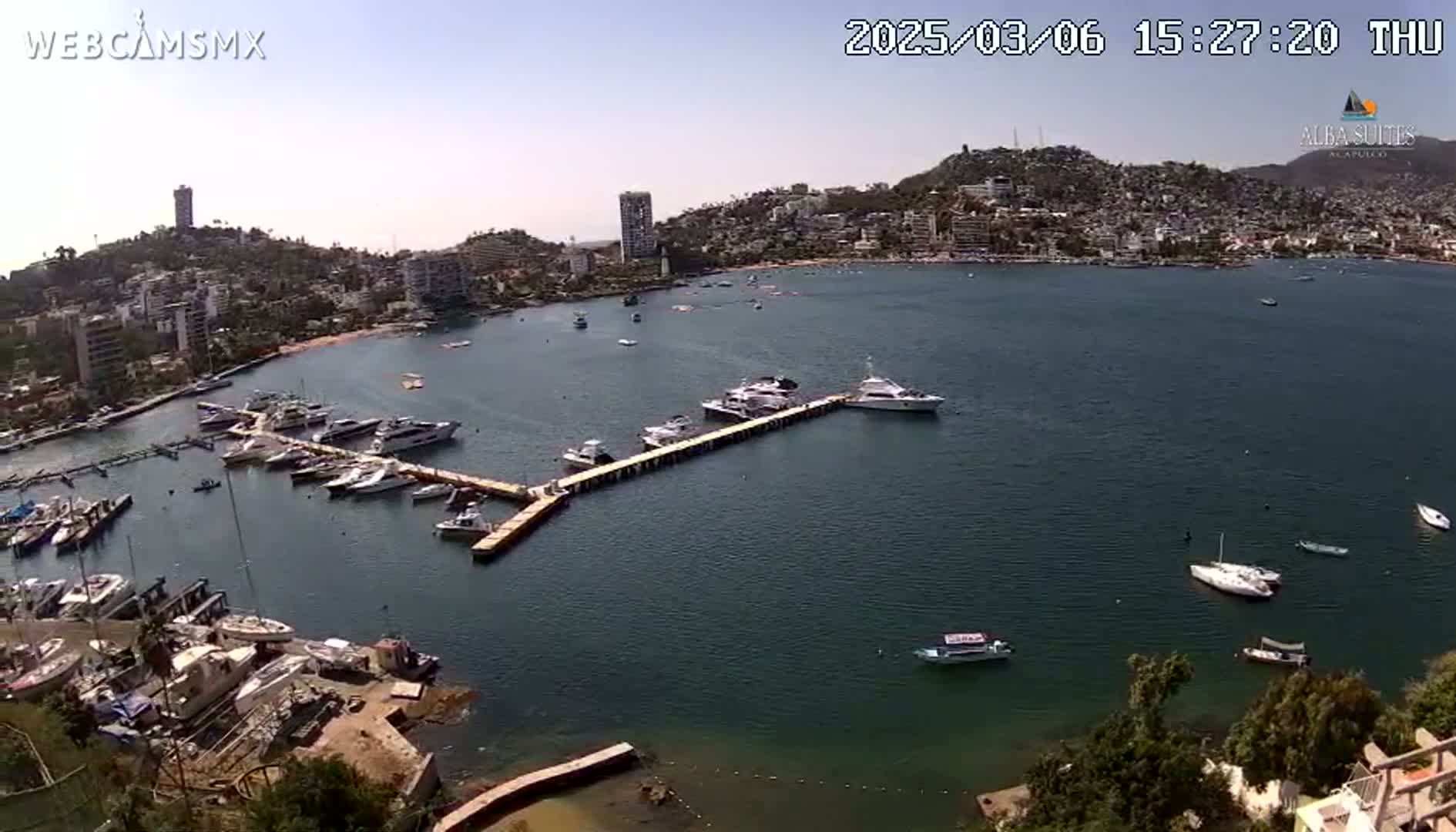 A marina filled with numerous boats and yachts is situated in a calm bay, with a hillside town visible in the background under a clear, sunny sky.