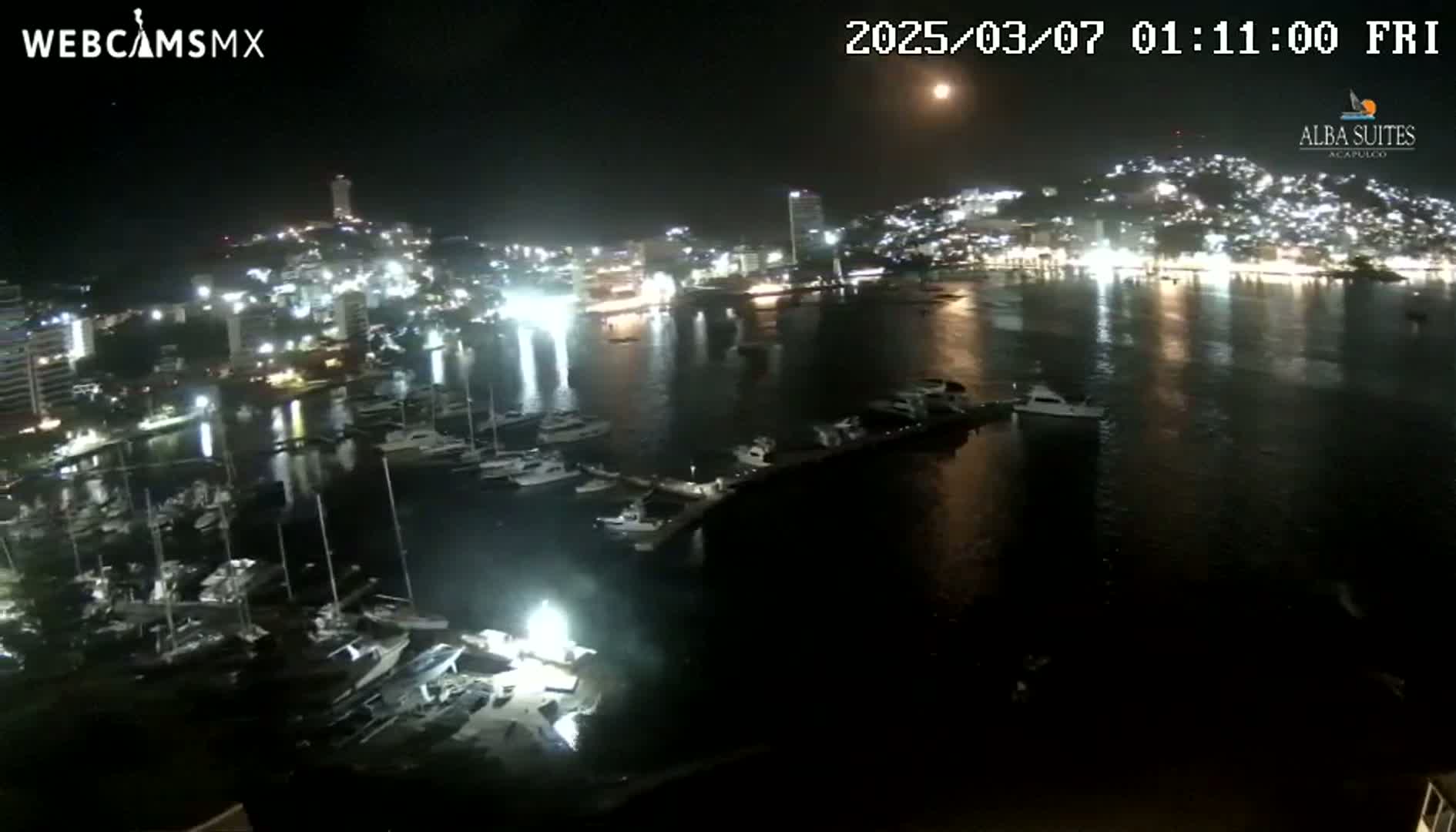 A nighttime aerial view of a harbor filled with boats, with a brightly lit city in the background under a mostly clear sky showing a bright moon.
