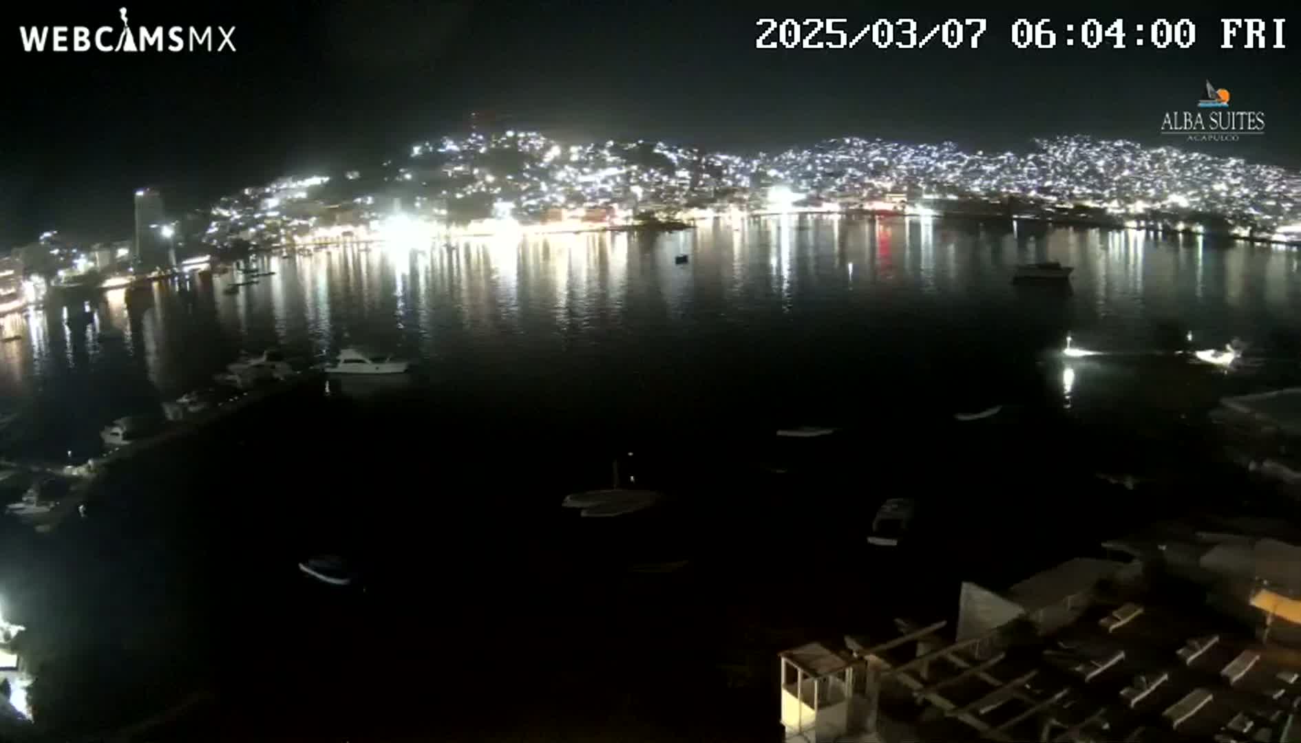 A nighttime view of a brightly lit city across a dark body of water, with several boats visible.