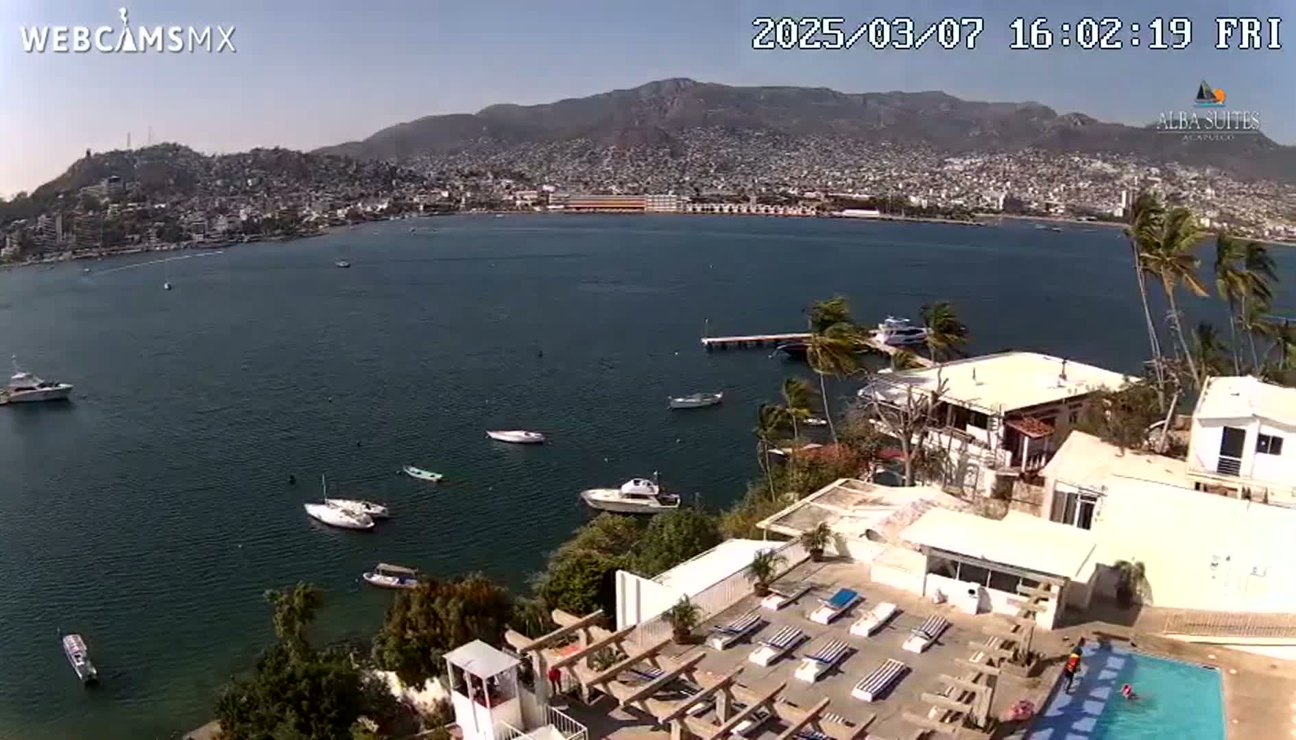 An aerial view shows a calm bay with several boats, a city nestled against mountains in the background, and a resort with a swimming pool in the foreground under a clear, sunny sky.