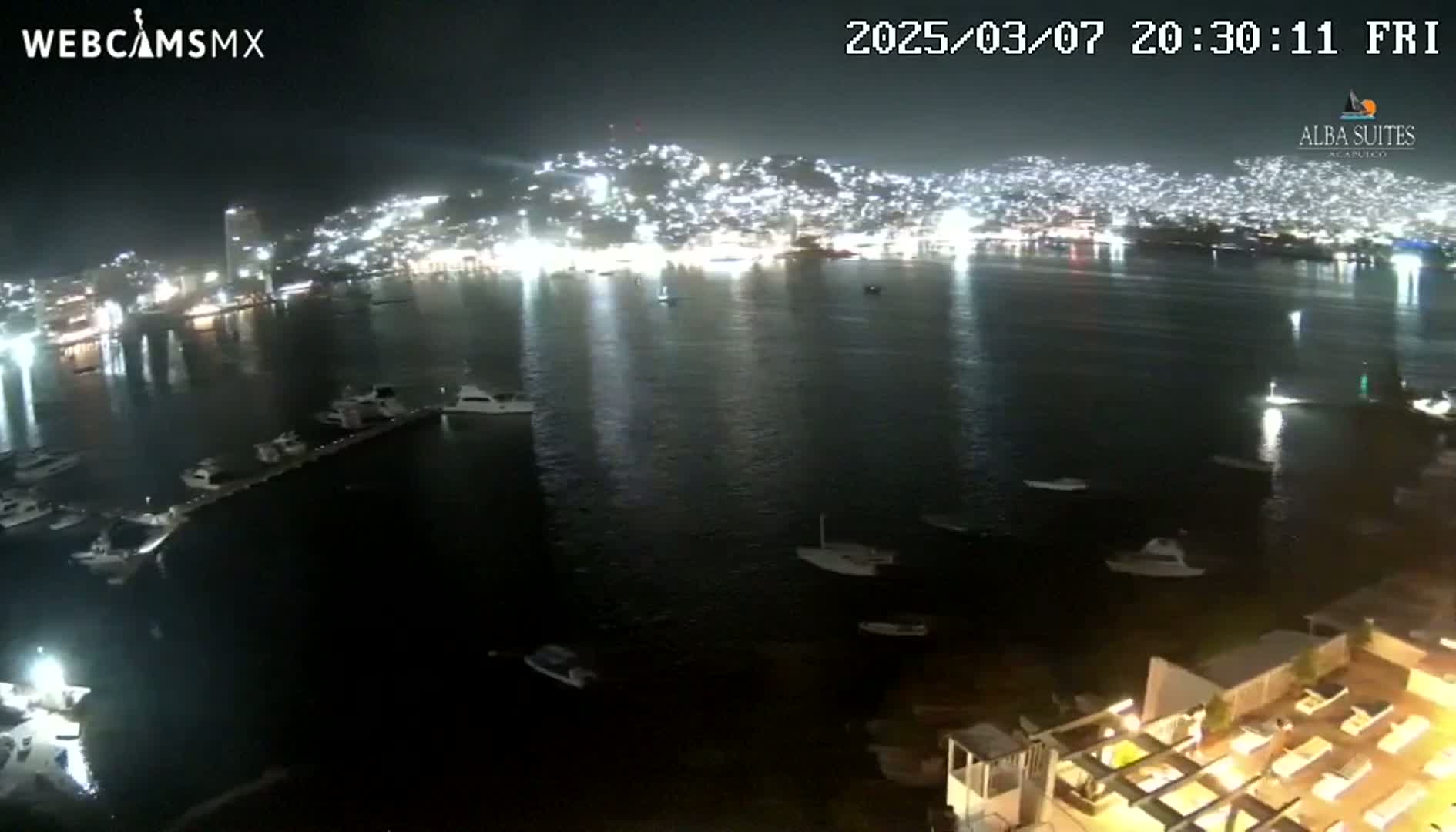 A nighttime view of a city illuminated along a dark body of water with numerous boats docked and afloat.