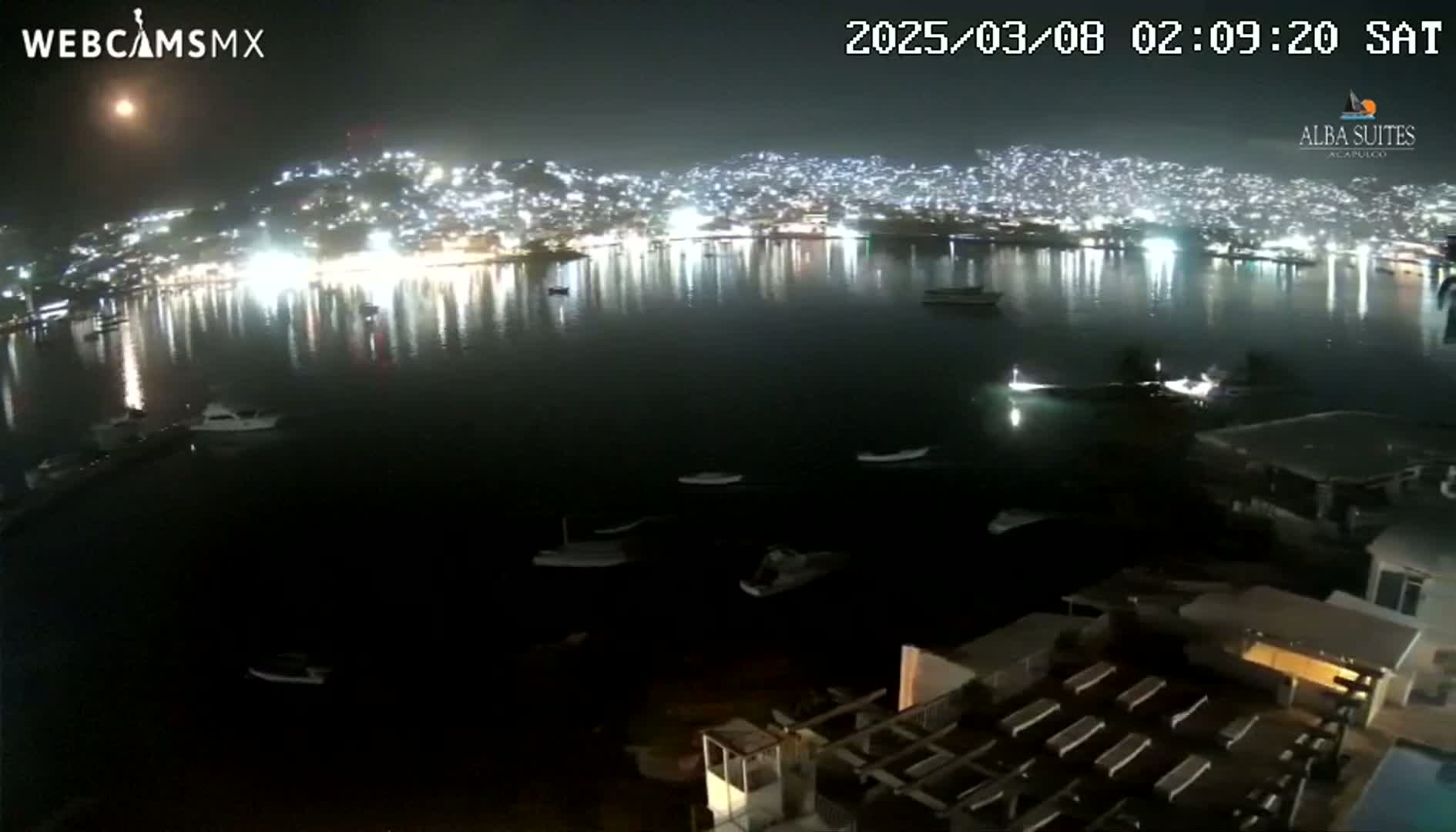 A nighttime view of a city illuminated along a calm body of water with several boats visible, under a slightly hazy sky.