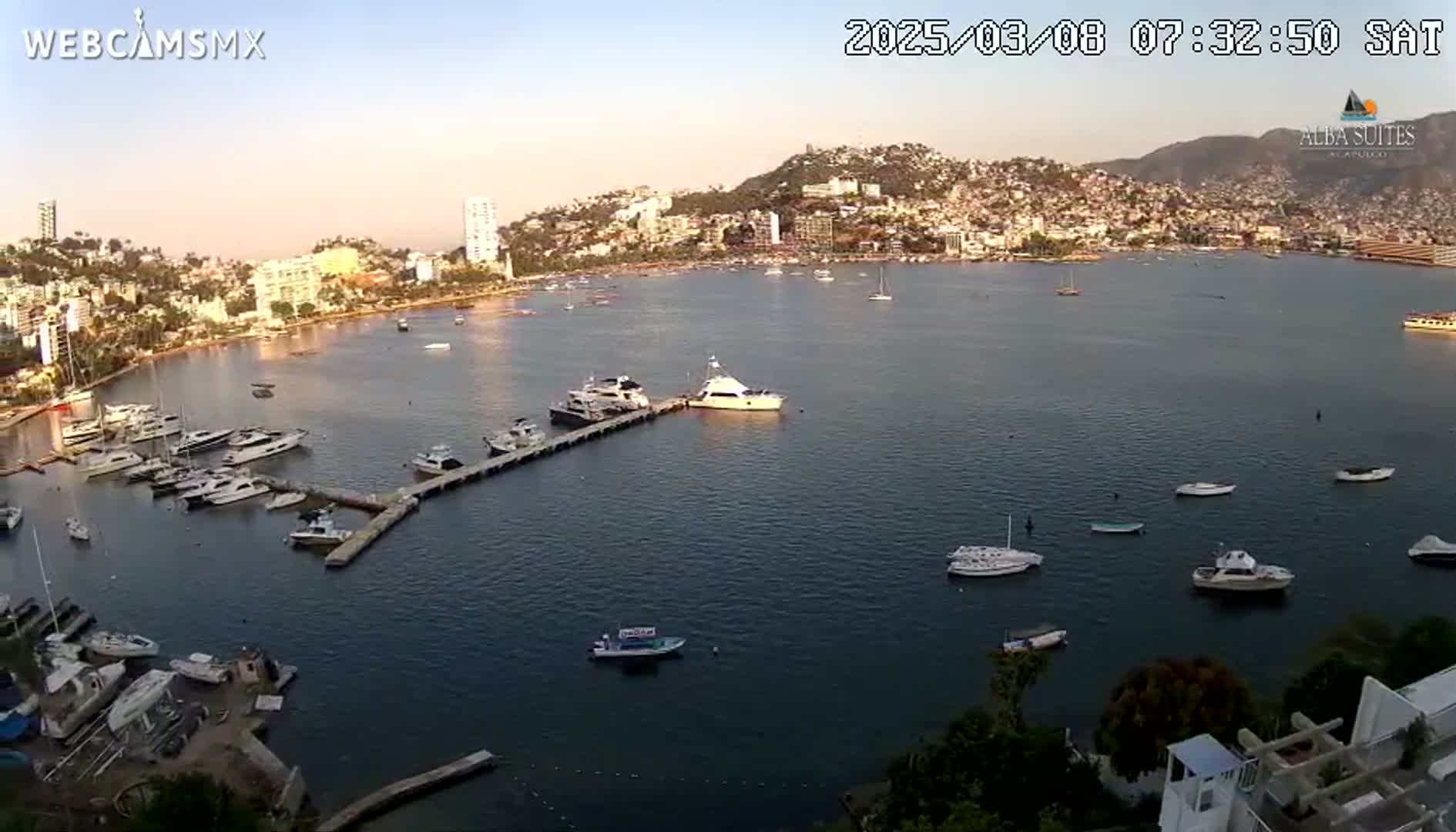 A calm bay with numerous boats and yachts in a marina, nestled beside a city built on a hillside under a clear, light sky.