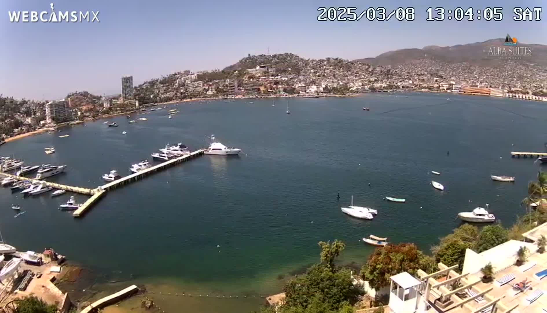 An aerial view of a calm bay with numerous boats docked at a pier, a town nestled on a hillside in the background under a clear, sunny sky.