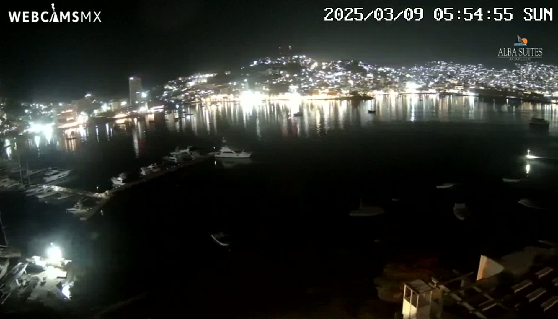 A nighttime view of a harbor filled with boats, reflecting the lights of a brightly lit city across the water.