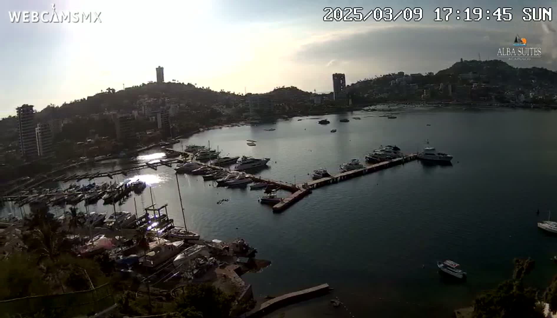 A marina filled with numerous boats is situated in a calm bay, surrounded by a hillside town under a mostly sunny sky with some clouds.
