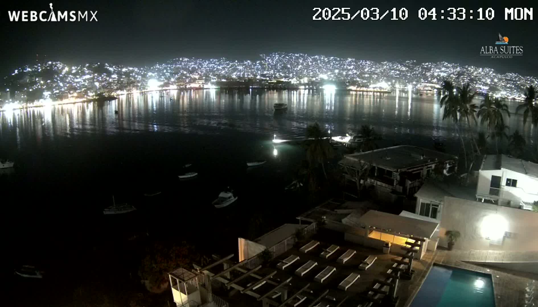 A nighttime view of a city illuminated across a bay, seen from a high vantage point overlooking a building with a pool and lounge chairs.
