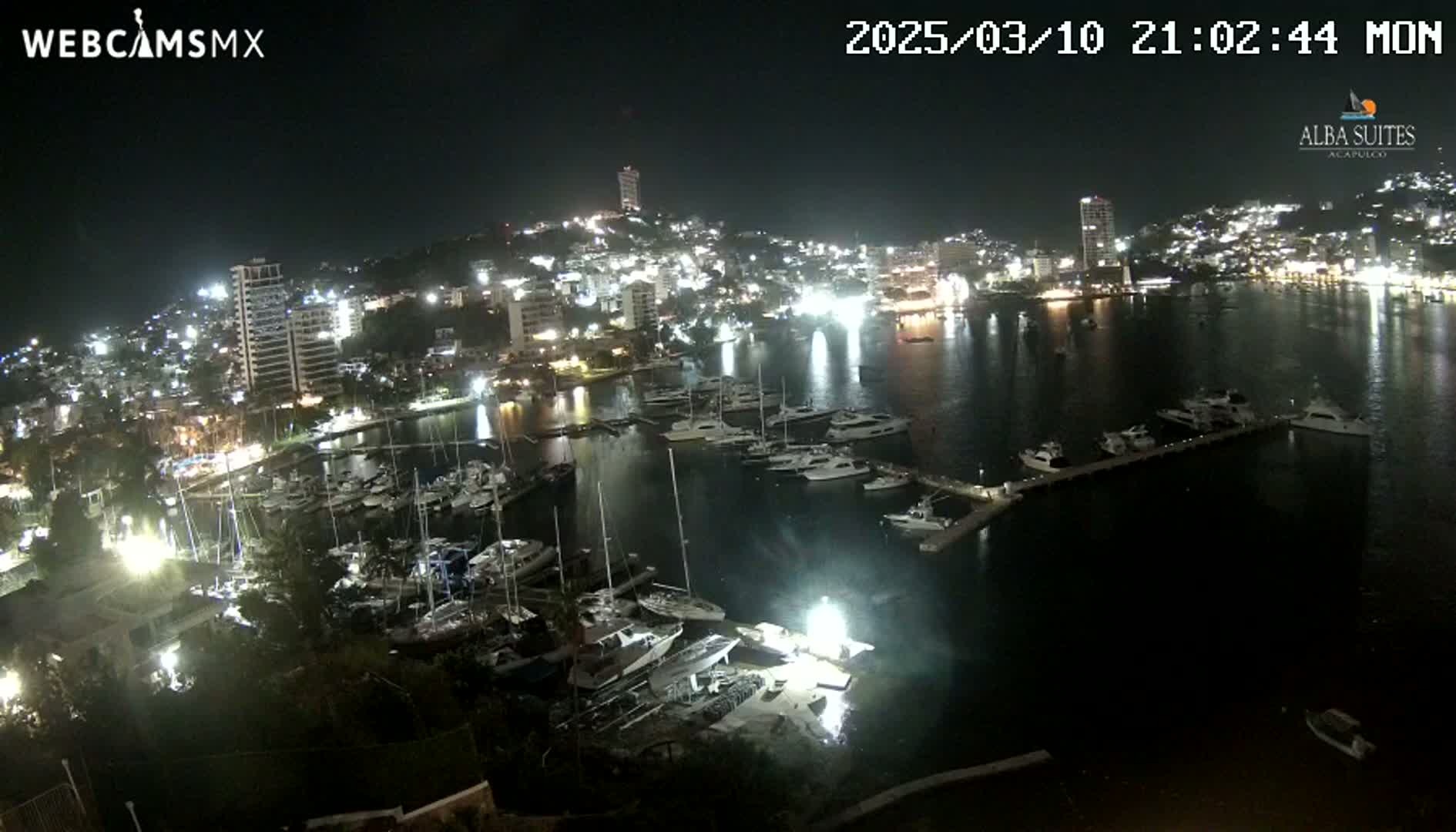 A nighttime aerial view of a harbor filled with boats, adjacent to a brightly lit city on a dark, clear night.