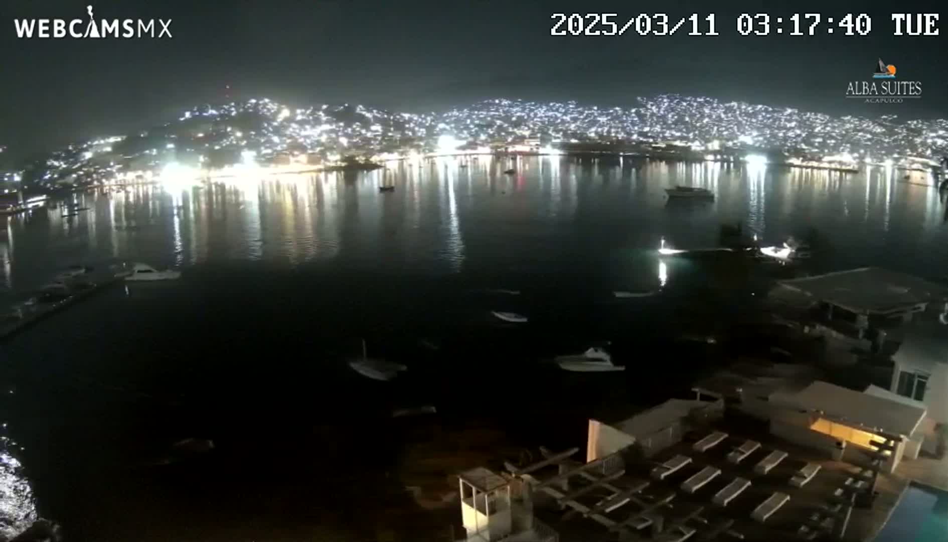 A nighttime view of a city illuminated across a calm bay, with several boats visible on the water and a building with outdoor seating in the foreground.