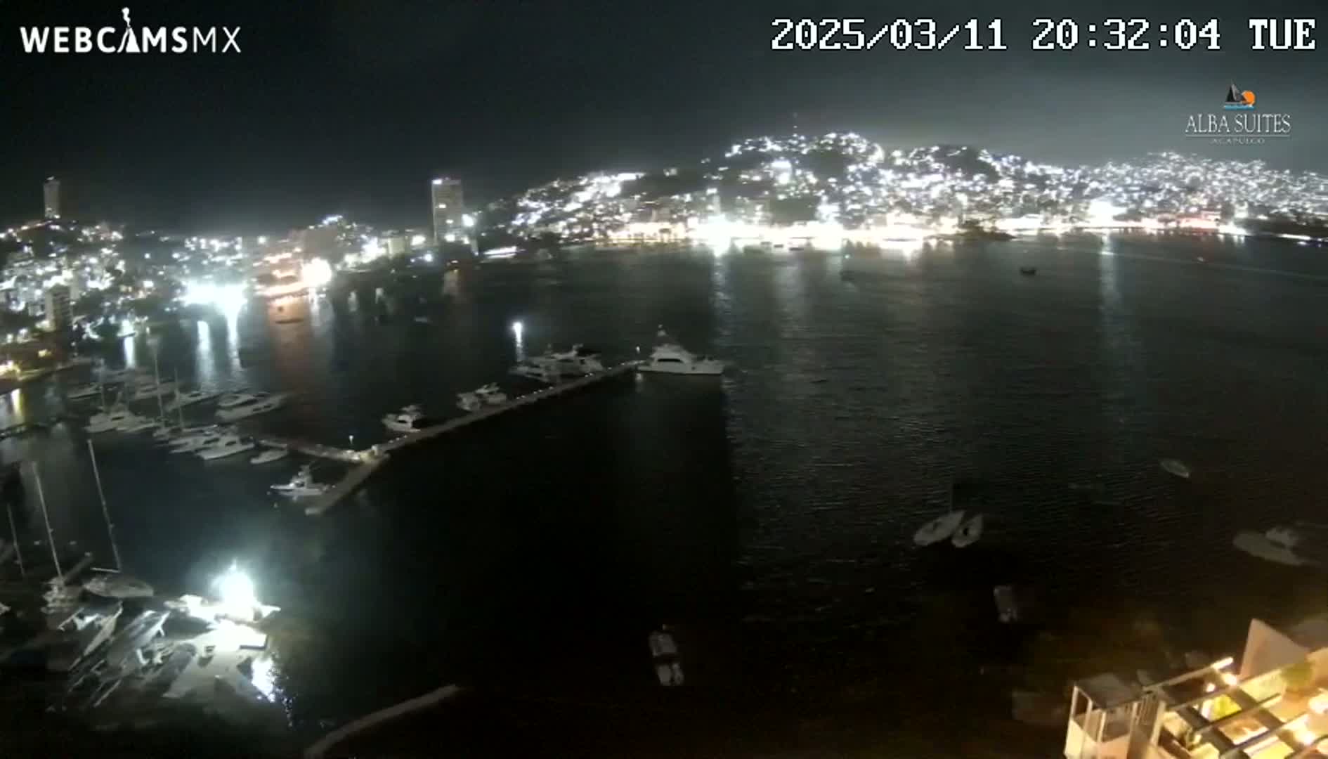 A nighttime, aerial view shows a harbor filled with boats, brightly lit buildings on a hillside across the water, and a calm bay.
