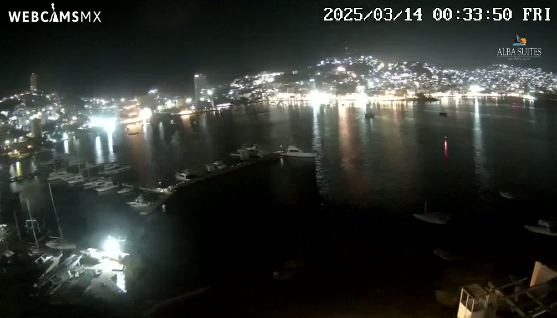 A nighttime, aerial view shows a harbor filled with boats, reflecting the city lights of a brightly lit town across the water.