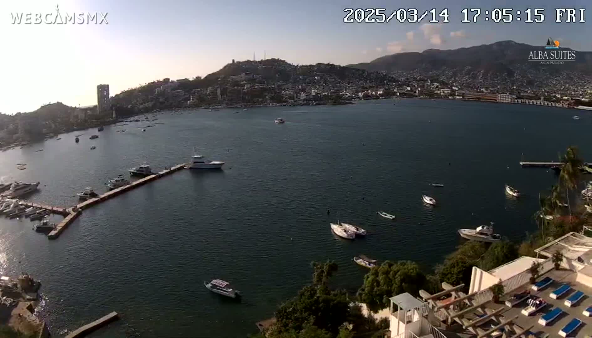 An aerial view shows a calm bay with numerous boats and a city nestled against hills under a clear, sunny sky.