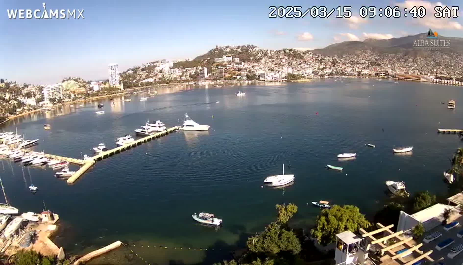 A calm bay with numerous boats docked at a marina, overlooks a city nestled against a hillside under a clear, sunny sky.