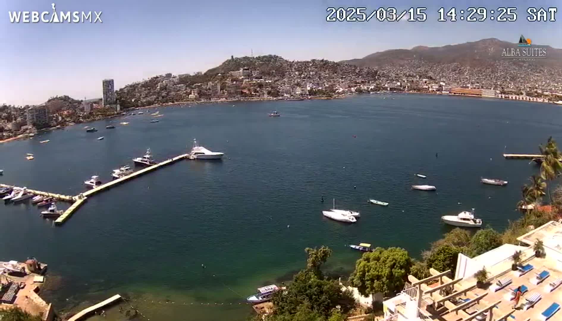 An aerial view shows a calm bay with numerous boats docked at a marina, near a hillside town under a clear, sunny sky.