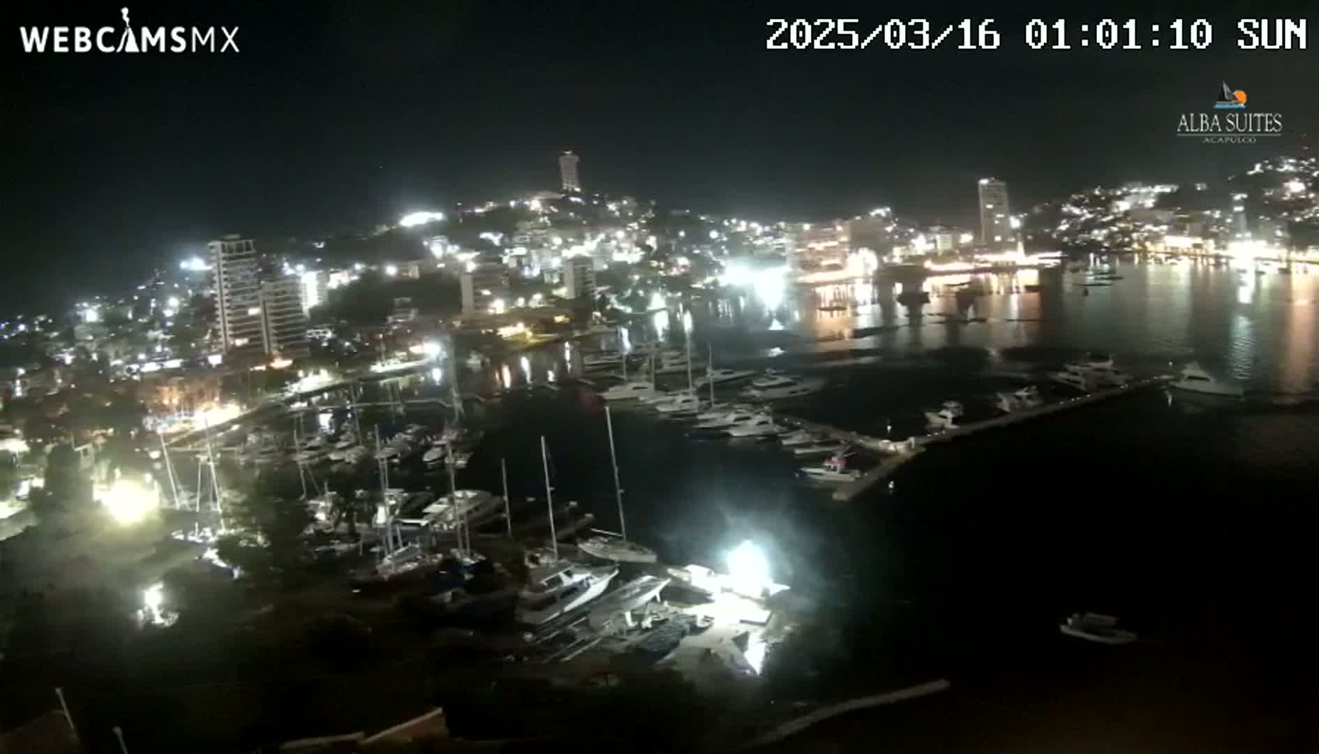 A nighttime aerial view shows a city marina filled with boats, illuminated buildings on a hillside, and calm water under a clear sky.