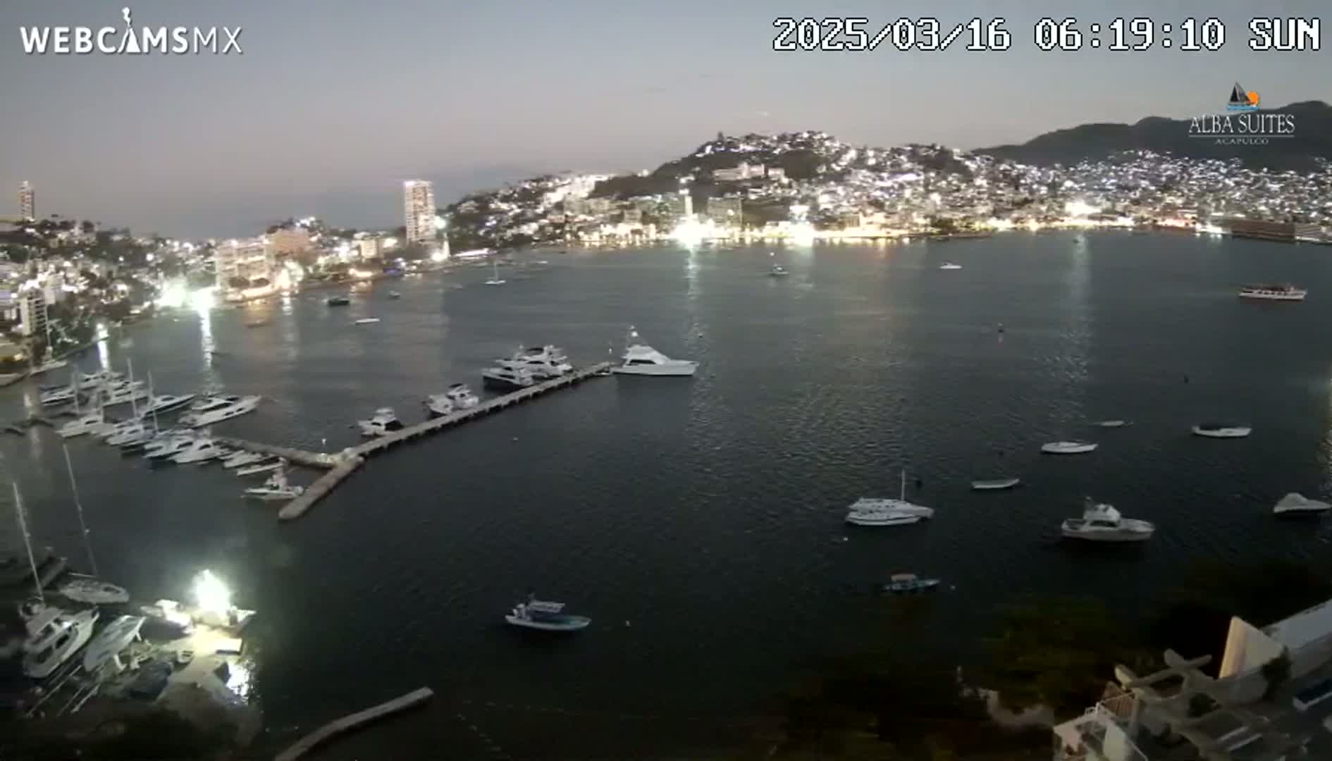 A nighttime, high-angle view shows a calm bay with many boats docked and anchored near a brightly lit city on a hill.