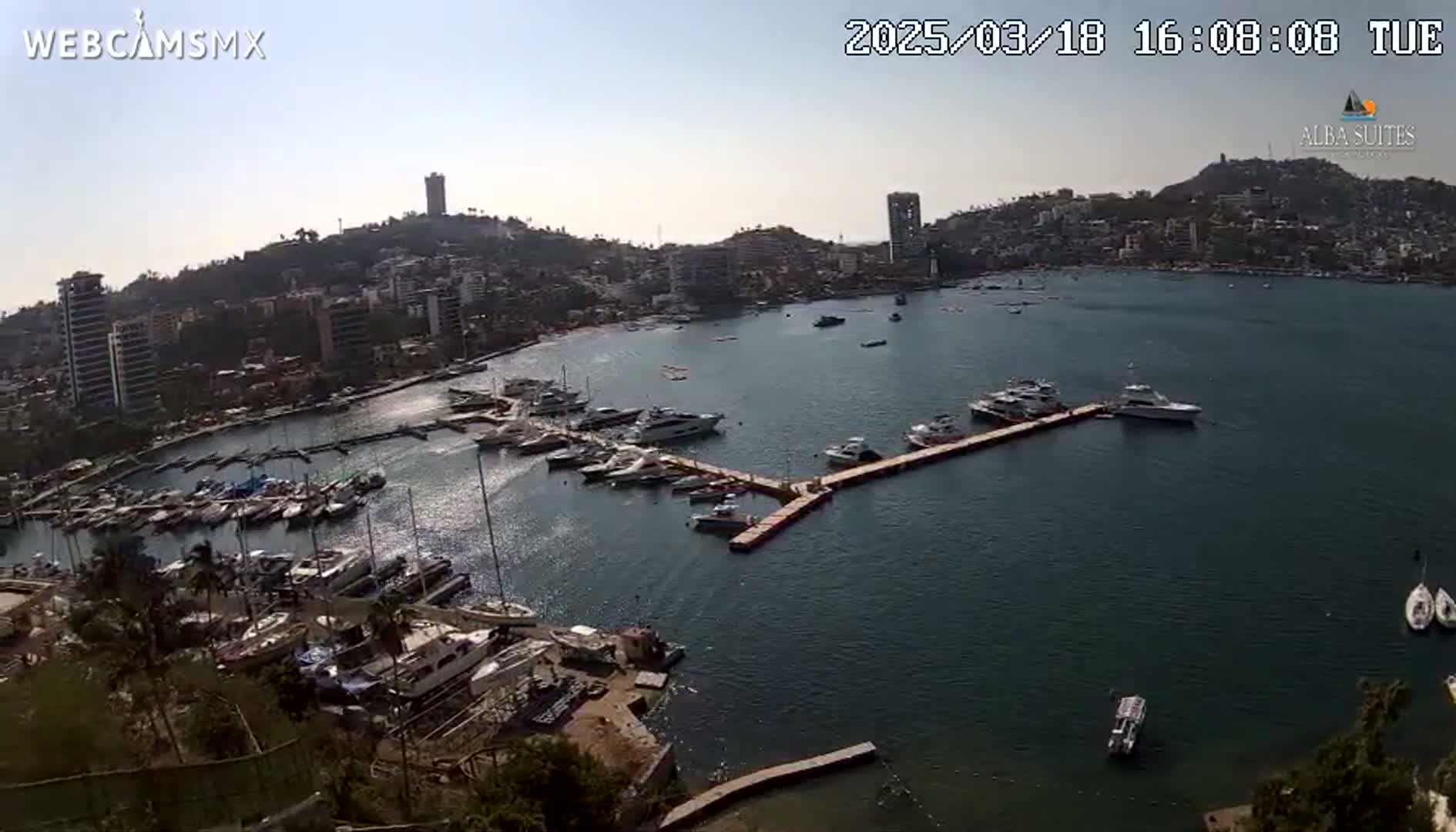 An aerial view of a marina filled with numerous boats, docked alongside a city built into a hillside, under a clear, sunny sky.