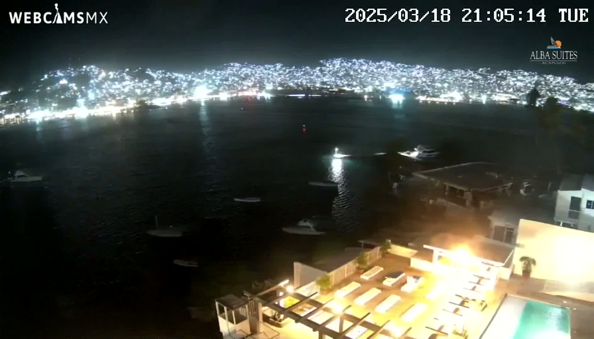 A nighttime view of a city across a dark body of water, with numerous boats visible, and a lighted patio area with lounge chairs and a pool in the foreground.