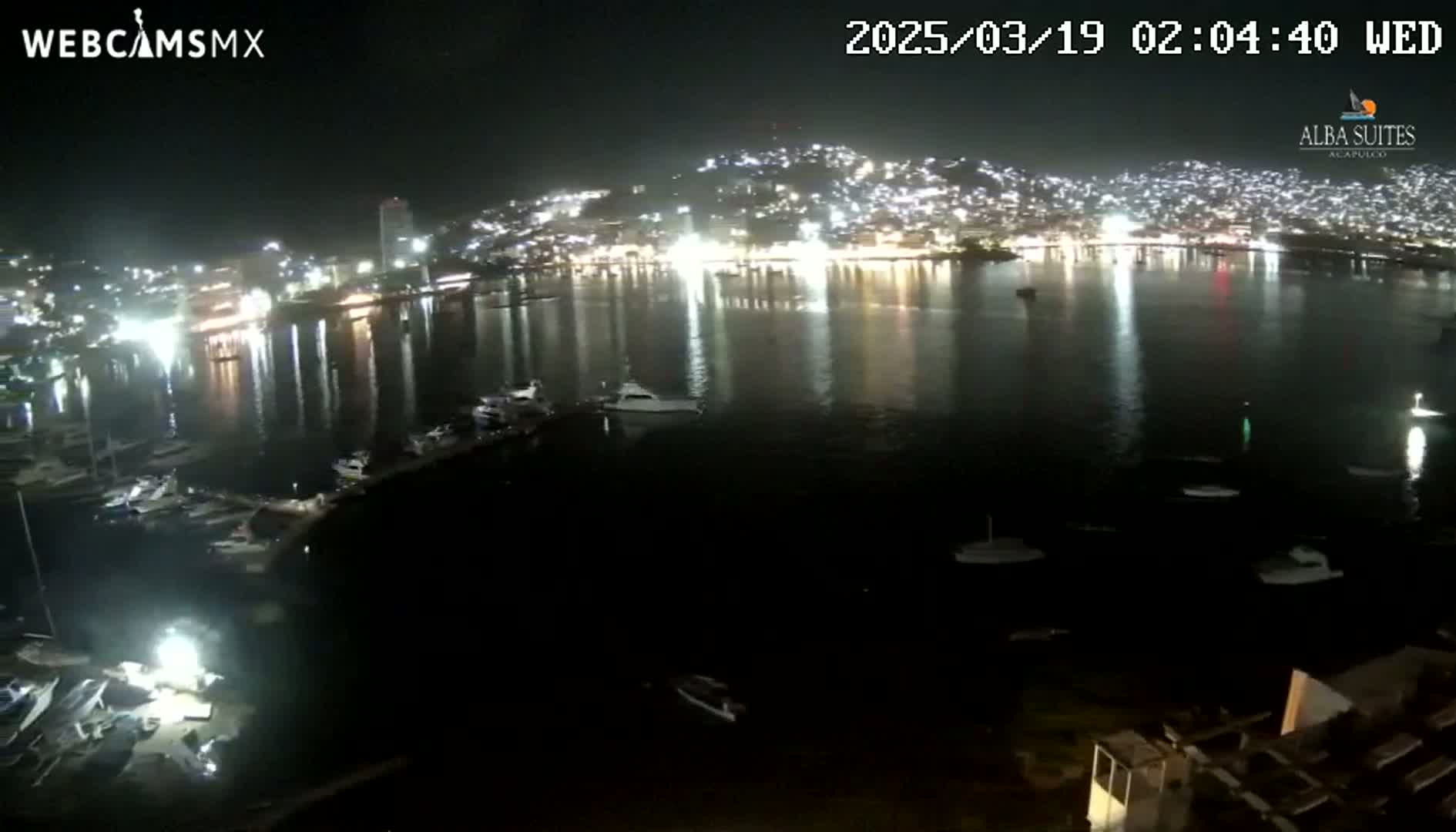 A nighttime view of a harbor filled with boats, reflecting the lights of a city built on a hillside.