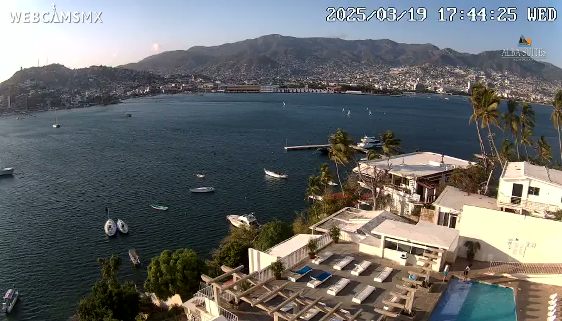 An aerial view shows a calm bay with several boats, a city nestled against mountains in the background, and a rooftop pool area in the foreground under a clear sky.