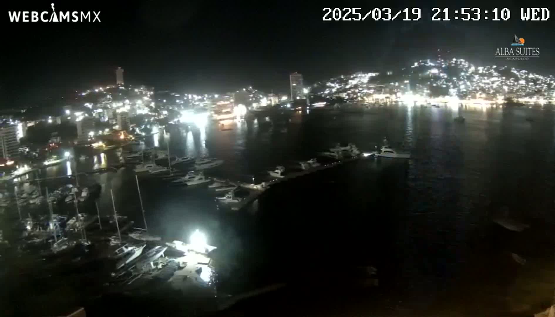 A nighttime aerial view shows a harbor filled with many boats, docked alongside a brightly lit city on a calm, dark body of water.