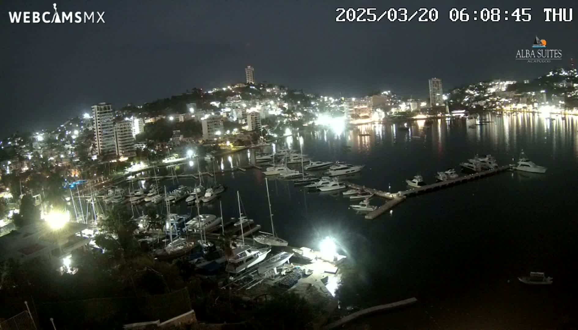 A nighttime view of a harbor filled with boats, with a city illuminated on a hill behind it under a dark sky.