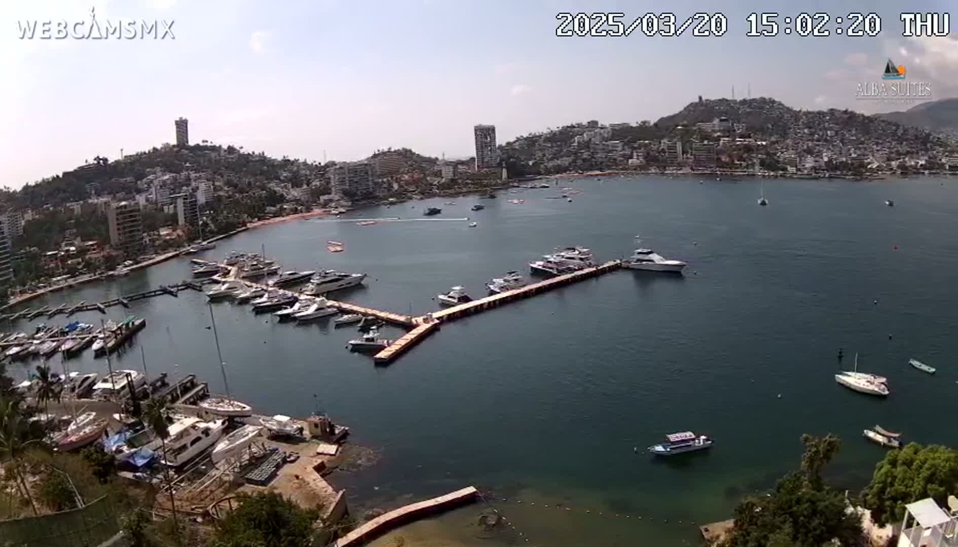 An aerial view shows a calm bay filled with numerous yachts and boats moored at a marina, nestled beside a hillside town under a sunny sky.