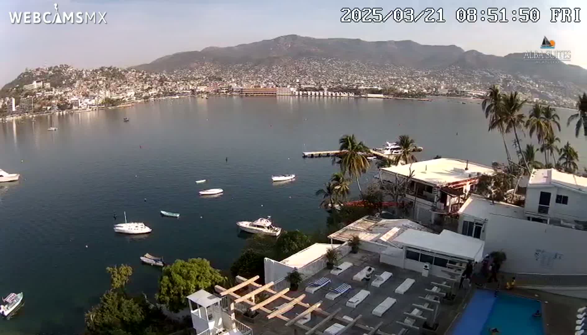 A calm bay with several boats, a city nestled against mountains in the background, and a hotel with a pool in the foreground under a clear sky.