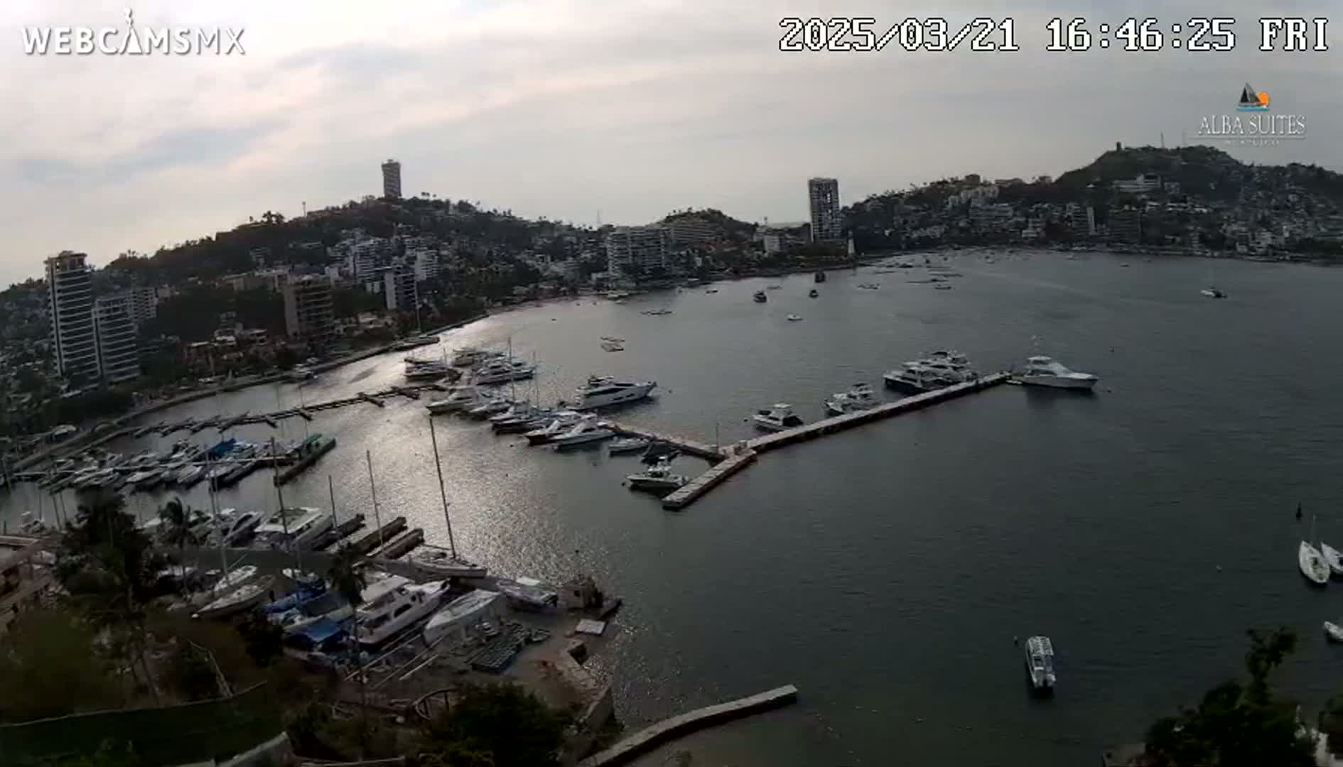 An aerial view shows a calm bay with numerous boats docked at a marina, surrounded by a city nestled against a hillside under an overcast sky.