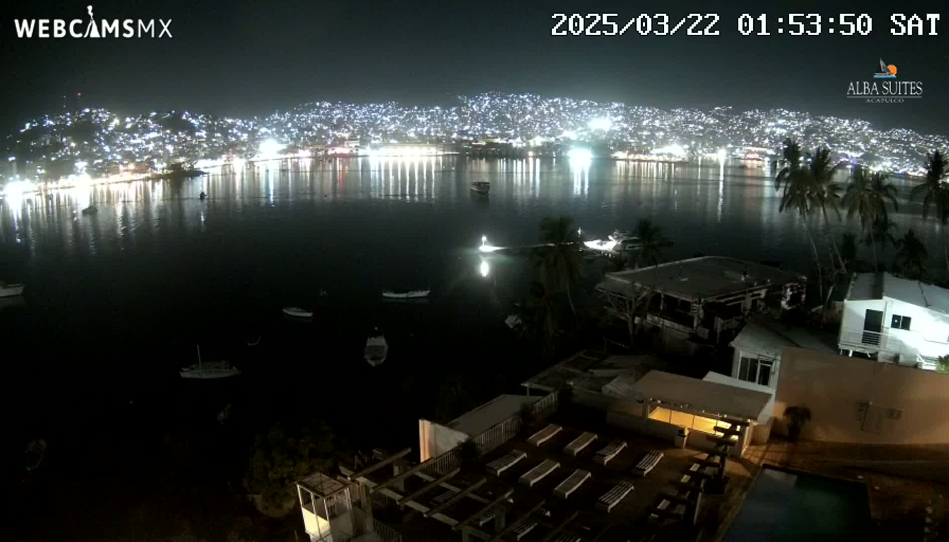 A nighttime view shows a city across a dark bay, with several boats on the water, and a building with a pool and sun loungers in the foreground.