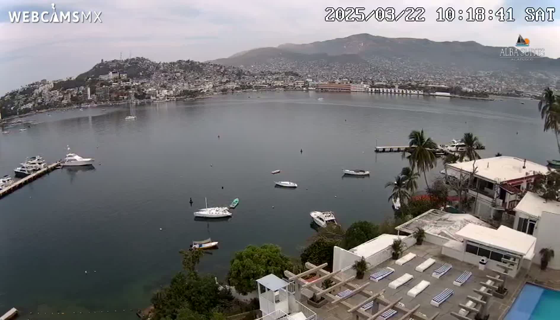 An overcast day reveals a harbor filled with boats, a city nestled against mountains in the background, and a white building with a rooftop pool and lounge chairs in the foreground.