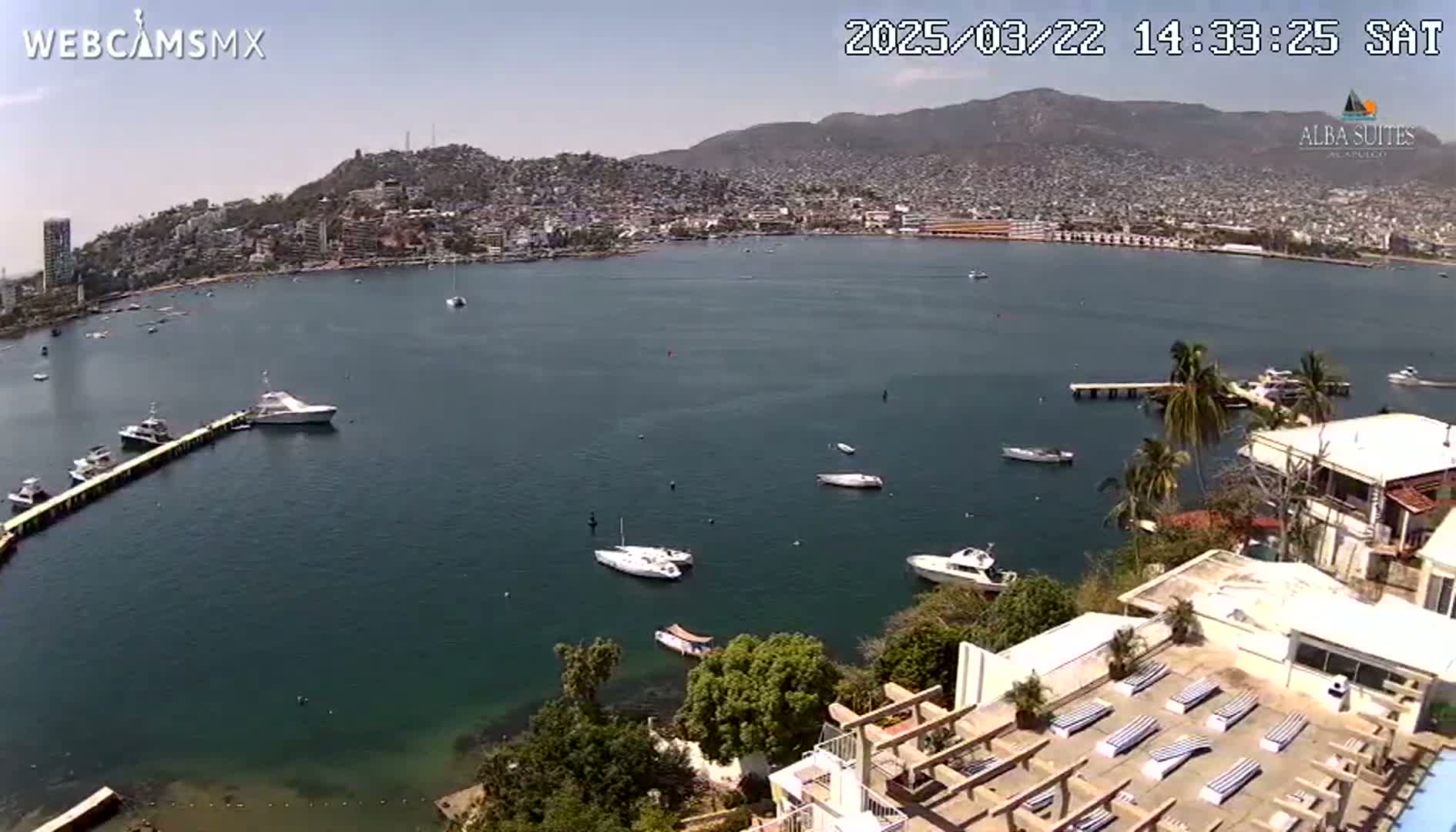 A sunny day overlooking a calm bay with numerous boats and a coastal city nestled against hills in the background.