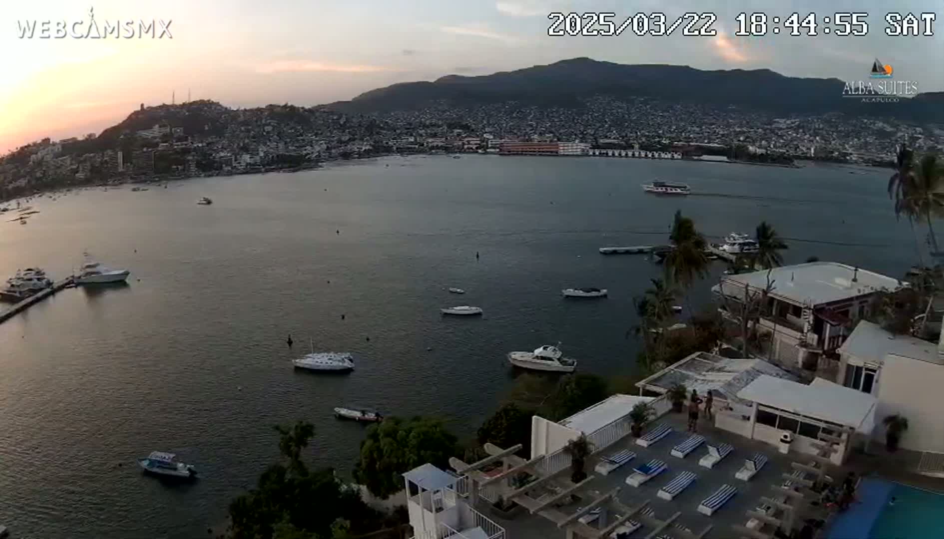 An aerial view of a calm bay with numerous boats, a coastal city at sunset, and a rooftop pool area with lounge chairs.