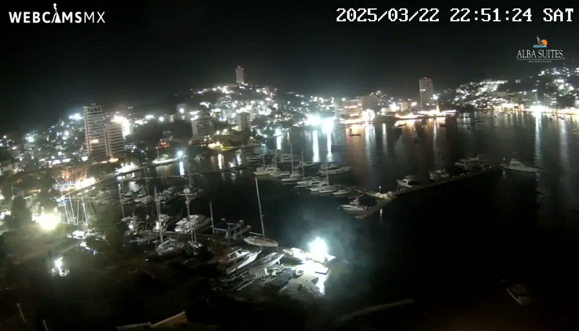 A nighttime view of a harbor filled with boats, adjacent to a brightly lit city on a calm, dark night.