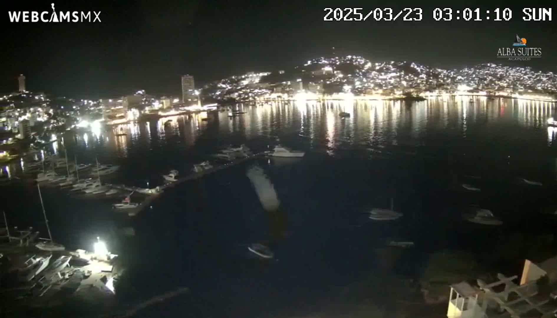 A nighttime view of a harbor filled with boats, with a brightly lit city rising on a hillside in the background.